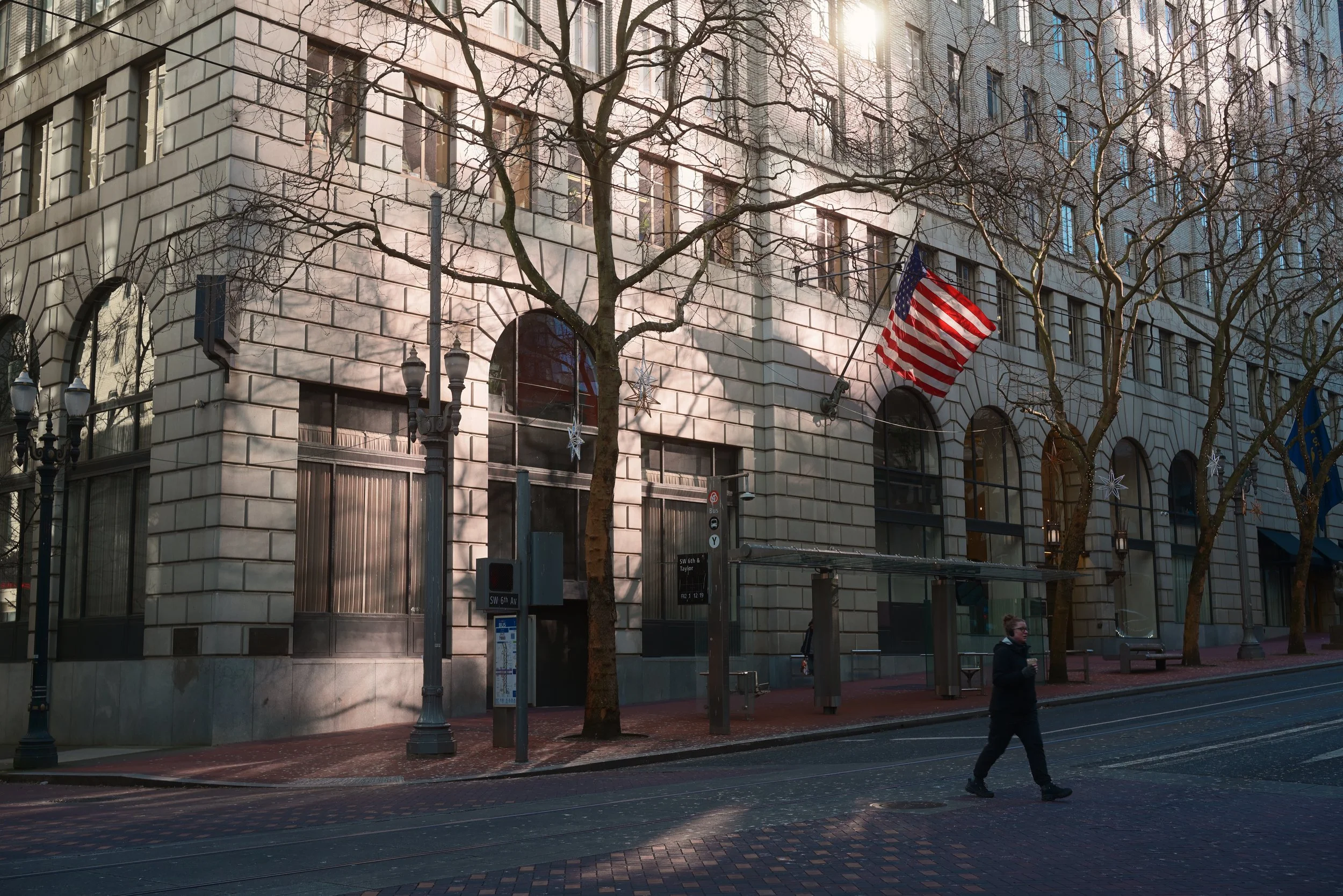 Street view of a building with American flag and holiday decorations, leafless trees, and a person walking on the sidewalk.