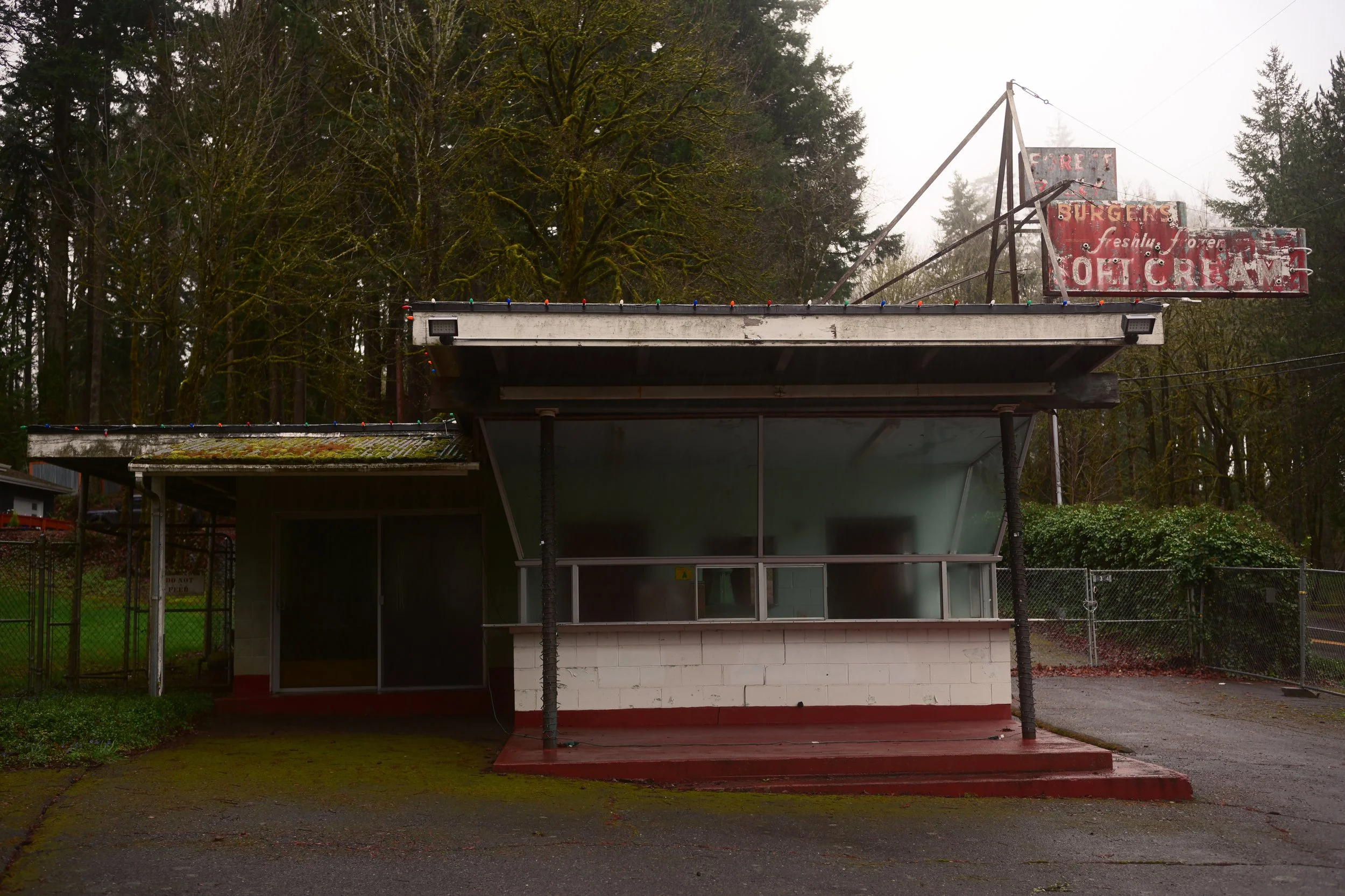 An old, weathered roadside food stand with a faded sign that reads 'Foster Burgers, Freshly Made, No Ice Cream.' The stand has a covered order window with a glass display and is surrounded by trees on a foggy day.