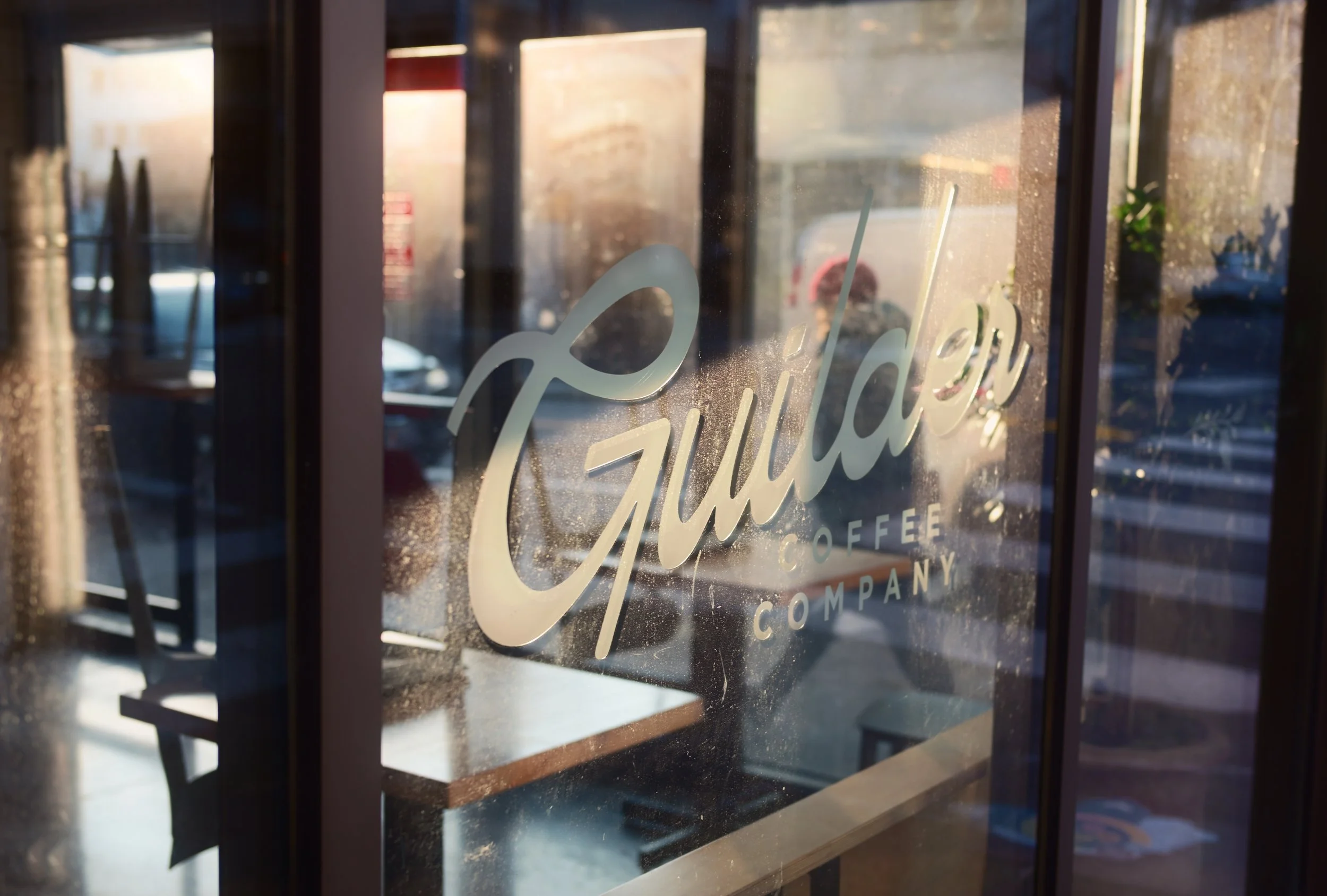 Close-up of a glass window with the logo 'Guilty Coffee Company' written on it, showing reflections of the street outside and interior seating