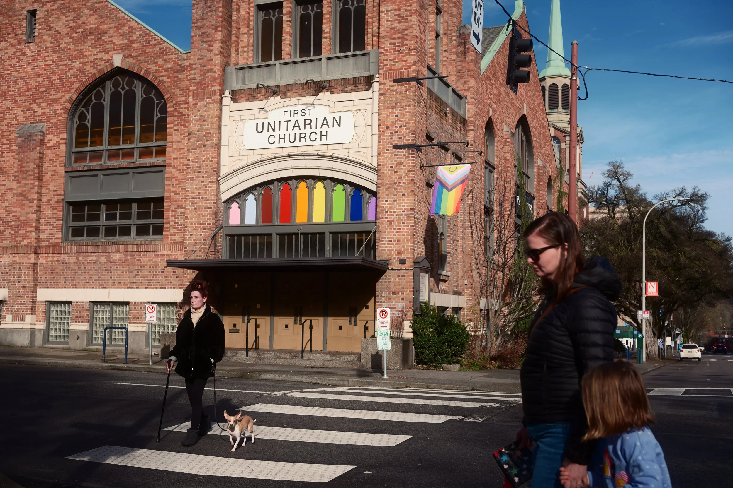 People crossing the street in front of a brick church with a sign that reads 'First Unitarian Church'. The church has rainbow-colored decorations and Pride flags displayed. One person is guiding a dog, while a woman and a girl walk hand-in-hand.