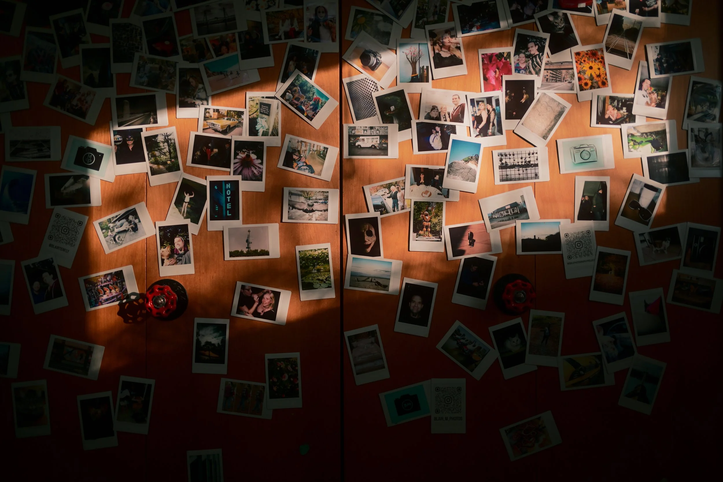 A wooden table covered with scattered printed photographs, some overlapping. The photos include various scenes such as people, landscapes, animals, objects, and QR codes. A couple of red objects and a small black device are also visible on the table.