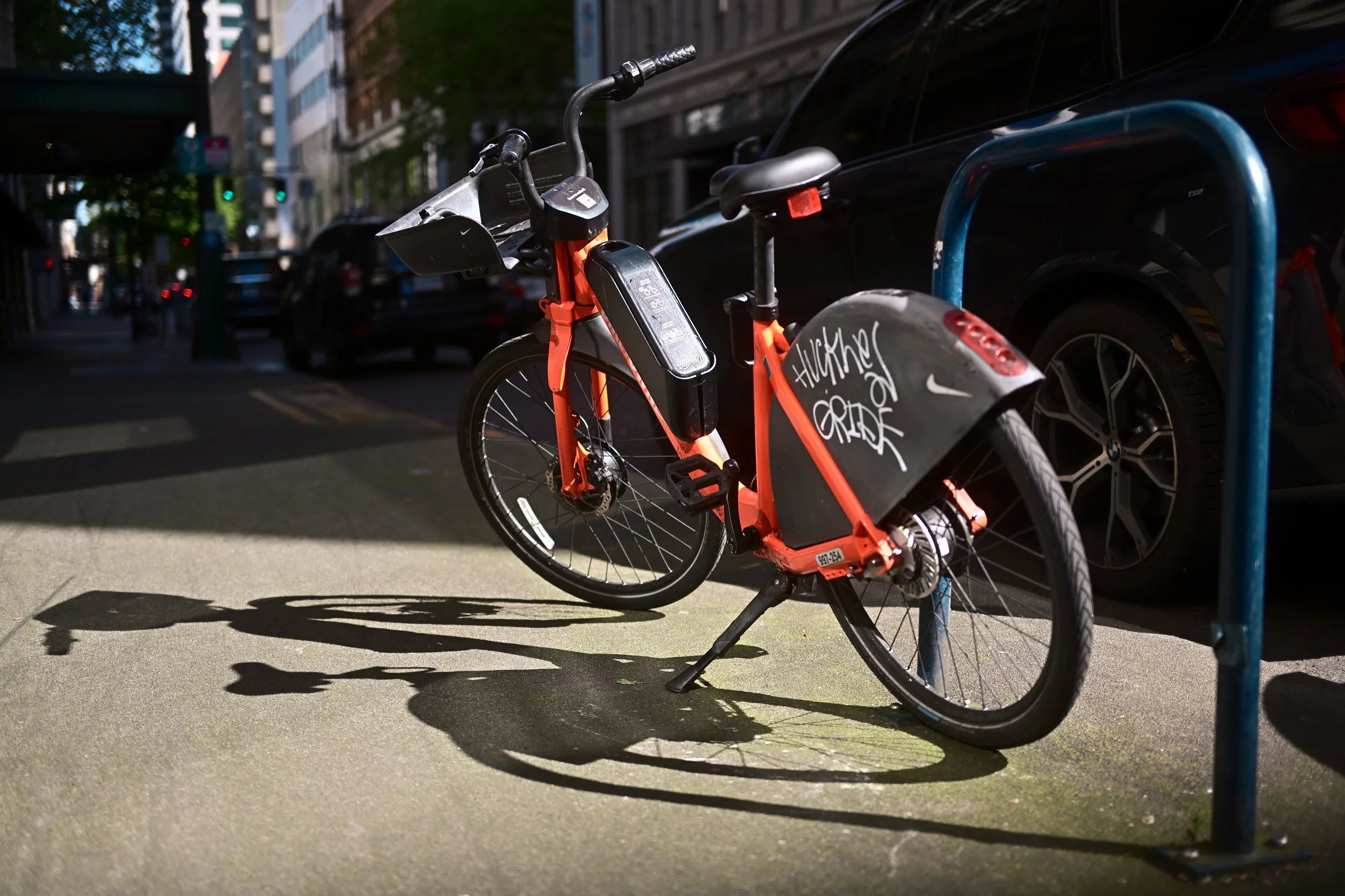 An orange bike parked at a bike rack on a city sidewalk with cars and buildings in the background.