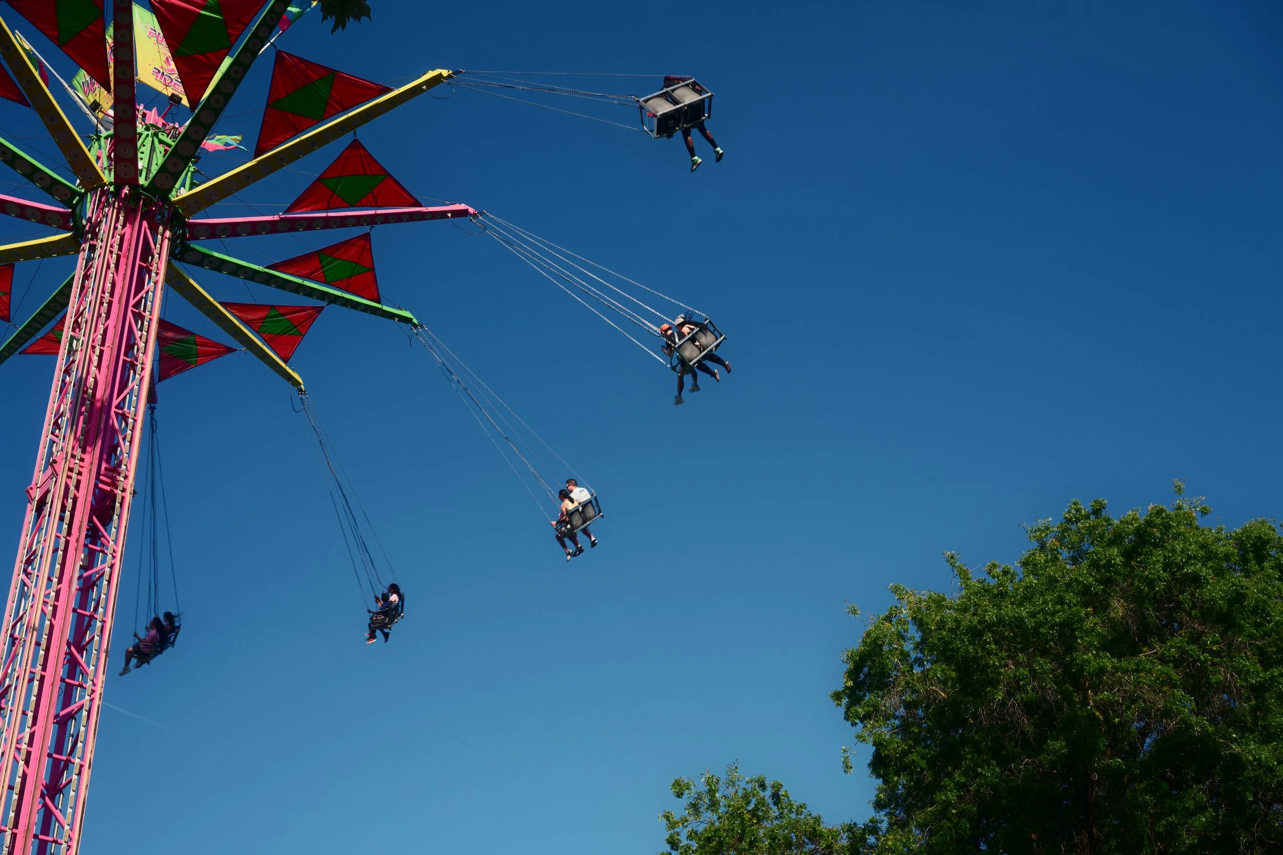 People riding on swings of a colorful ferris wheel against a clear blue sky, with green trees at the bottom right.