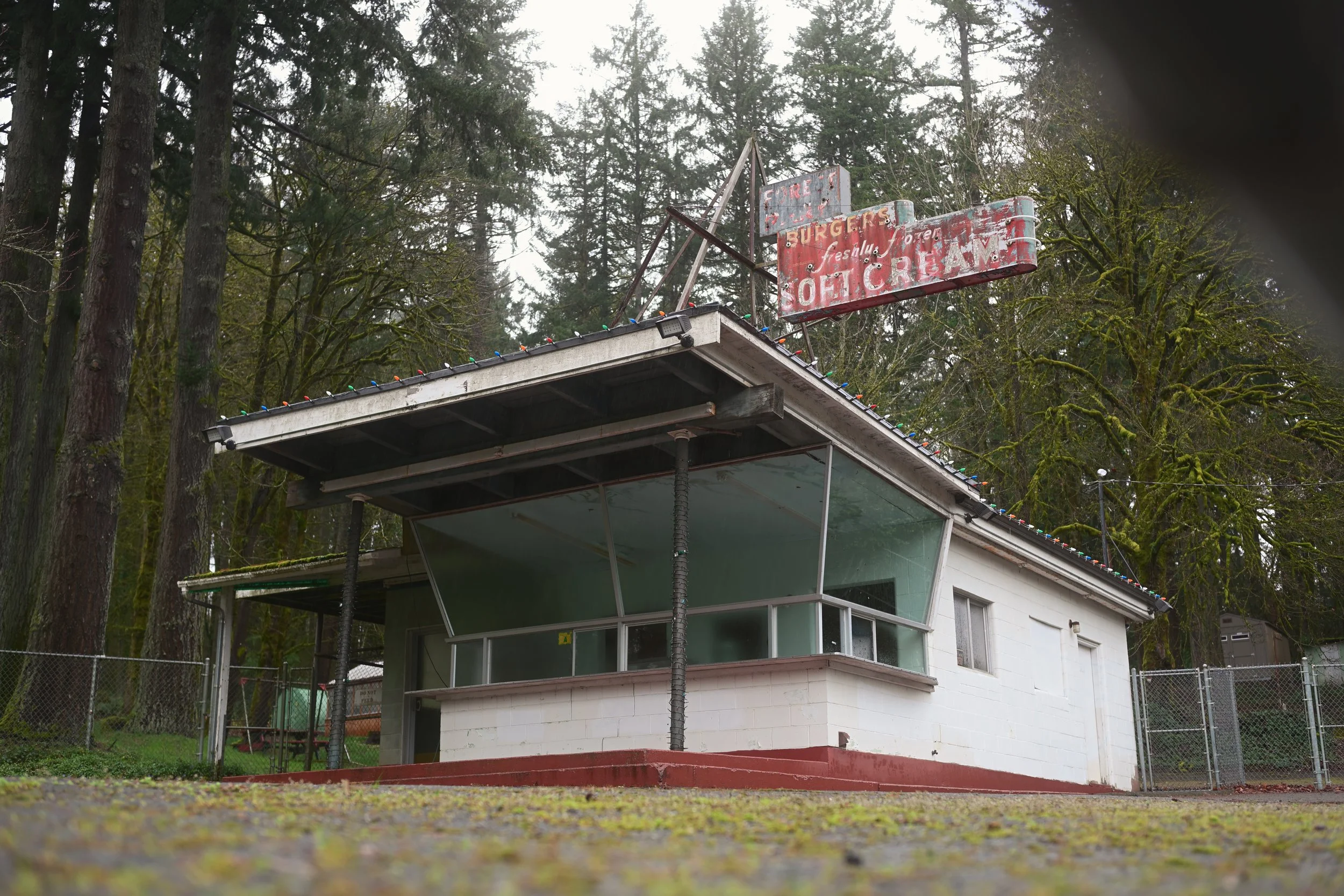 An abandoned roadside burger stand with a faded red and white sign that reads 'Burgers soft serve'. The structure has a glass-enclosed front and a small overhang decorated with Christmas lights, surrounded by tall evergreen trees.