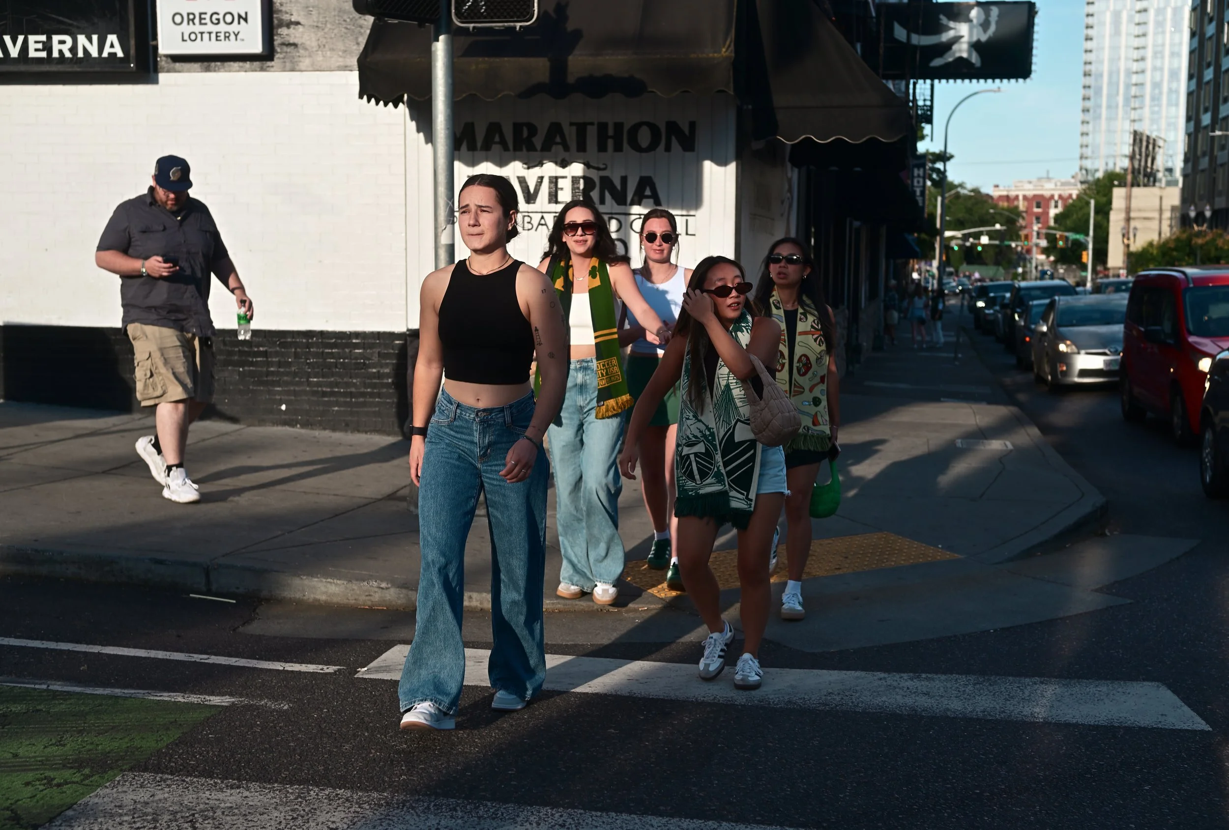 A group of five young women walking across a city street on a sunny day, with a man behind them looking at his phone.