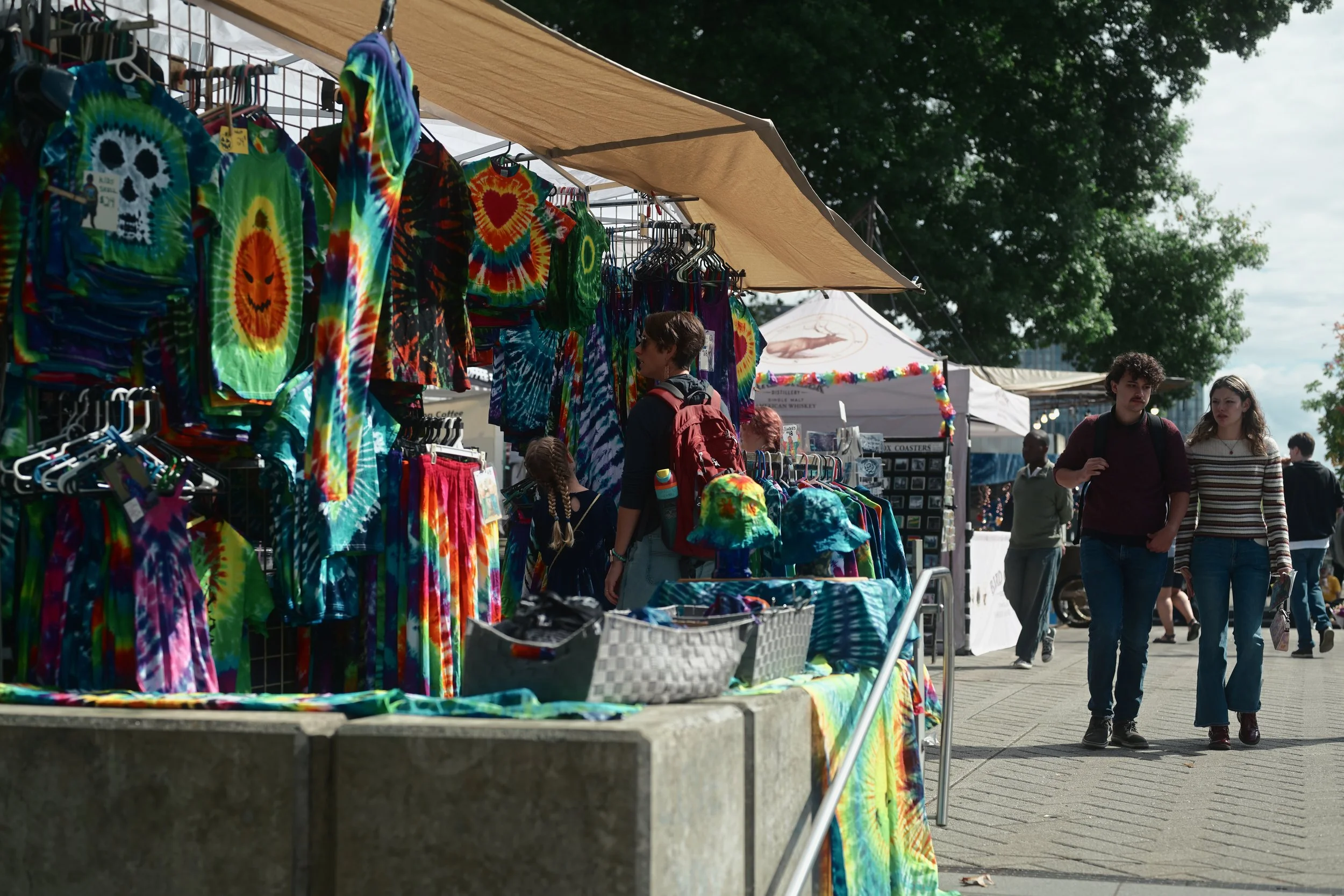 A street market stall displays colorful tie-dye shirts and clothing. People are shopping and walking in the background with trees and tents visible.