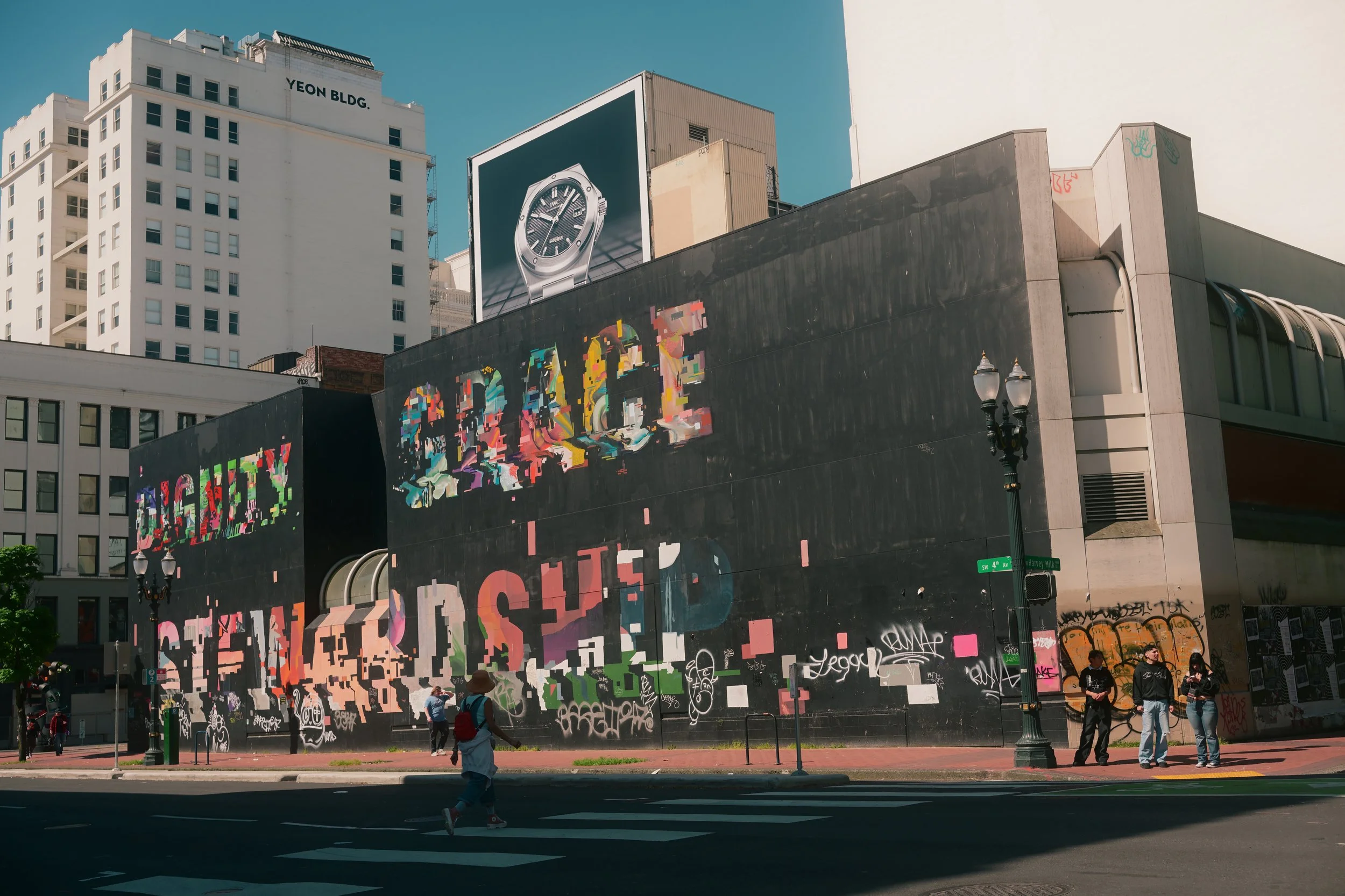 A city street scene with a large black building covered in colorful graffiti and digital art. People are walking in front of the building, and there are tall white buildings, billboards, and street lamps visible against a clear blue sky.