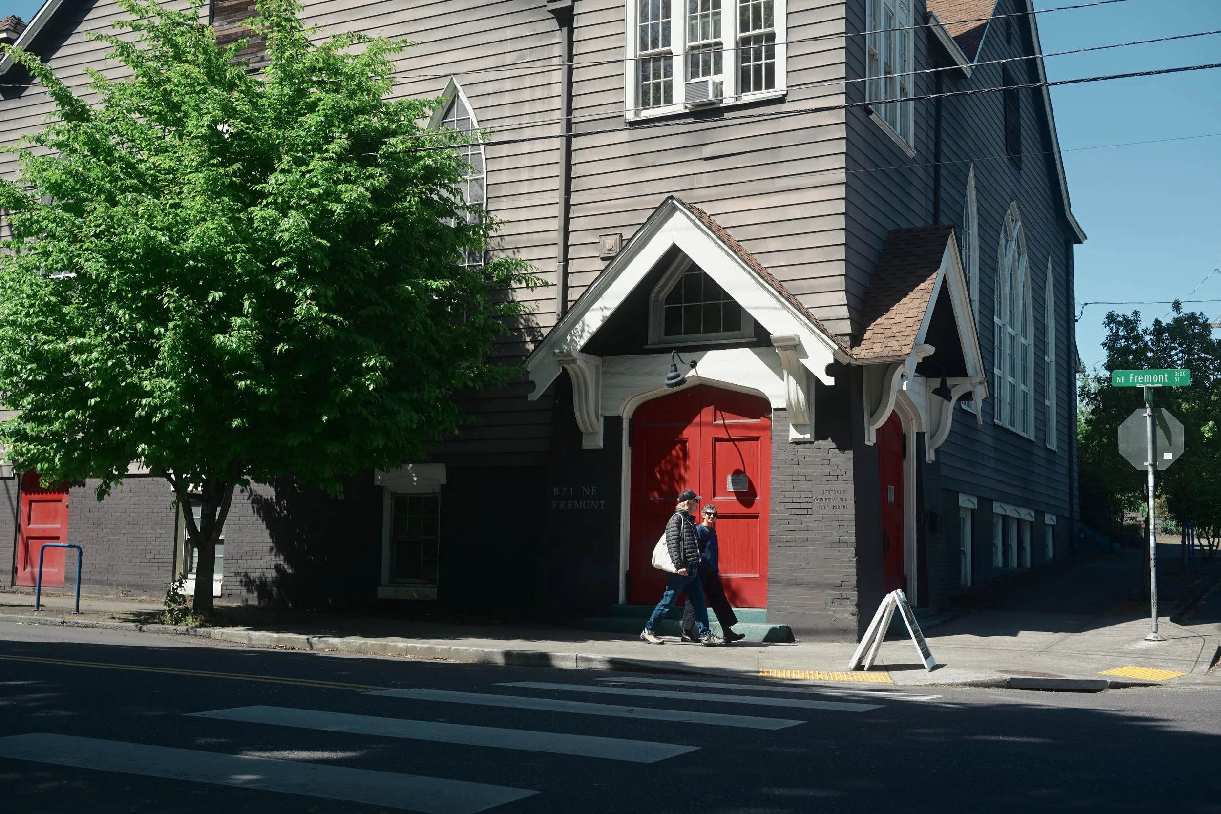 A large gray house with vertical siding and a steep gable roof with brown shingles, featuring a white trim and multiple large windows. The entrance has a red door with a white frame, and two people are walking past the house on the sidewalk. There are trees and a street sign that reads "NE Fremont" in the foreground.