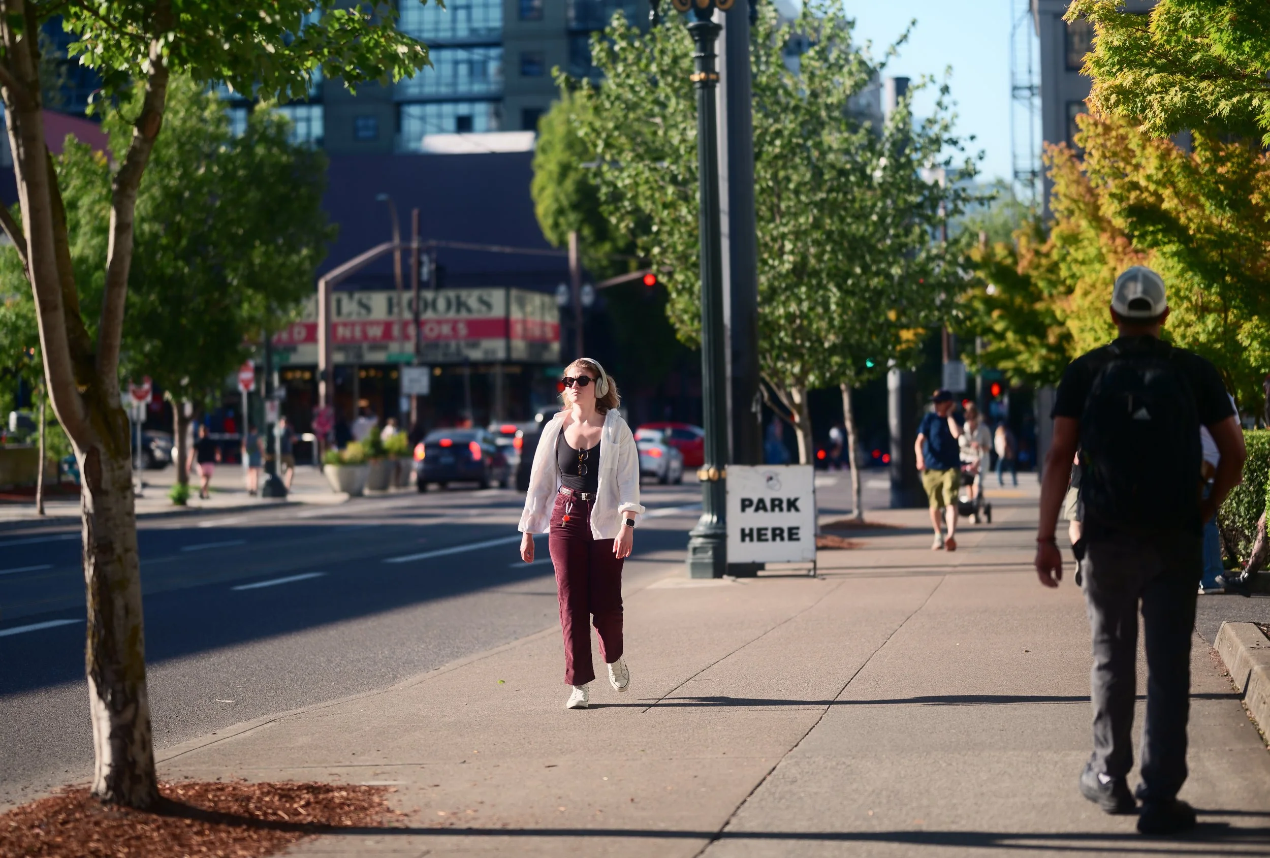 People walking on a sunny city sidewalk with green trees, cars, and buildings in the background. A woman with sunglasses, headphones, and a white jacket is in the center. A man with a backpack and cap walks away from the camera.