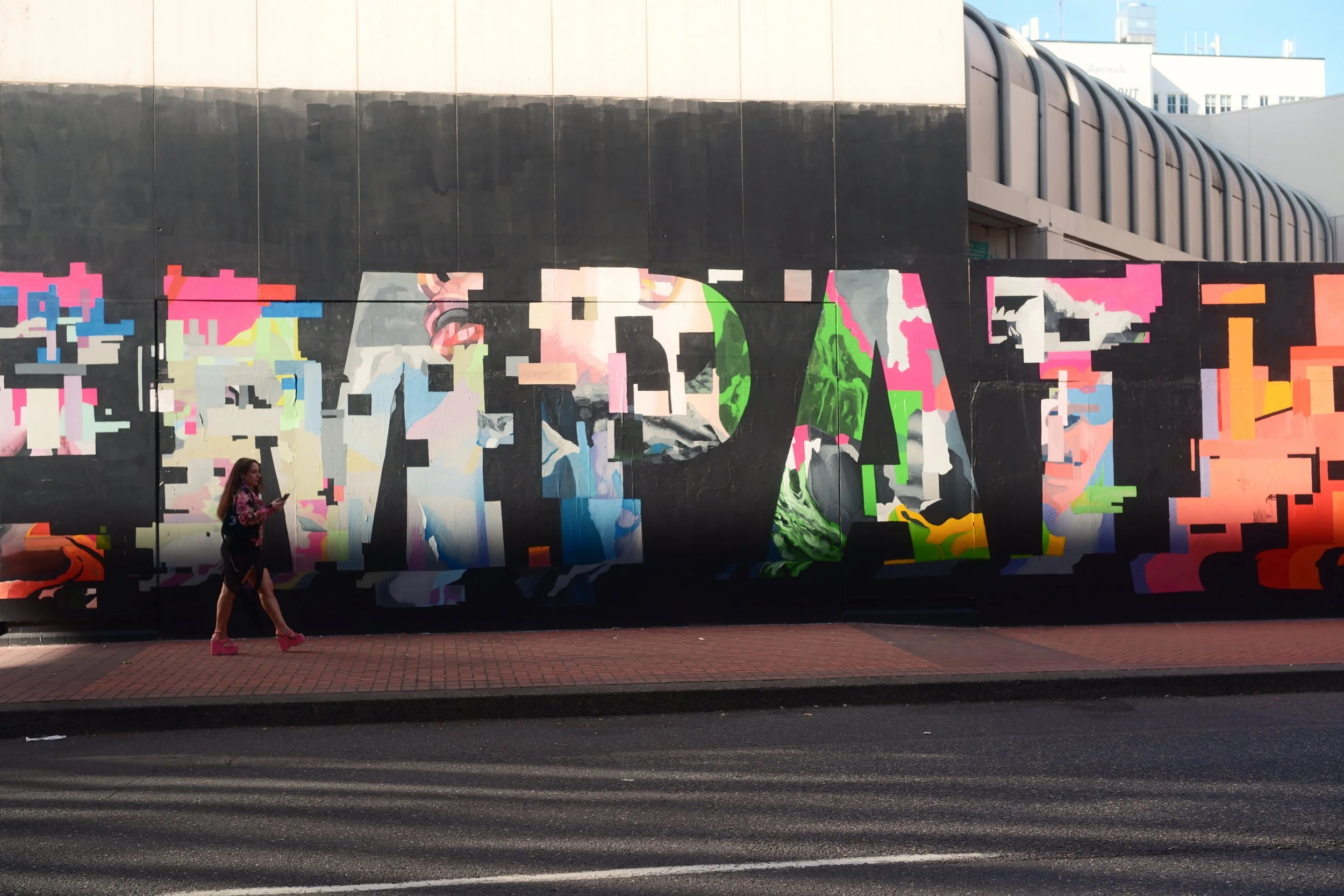A woman wearing pink shoes and a floral jacket walks past a long mural with colorful, abstract pixel art and a large face on a city street.