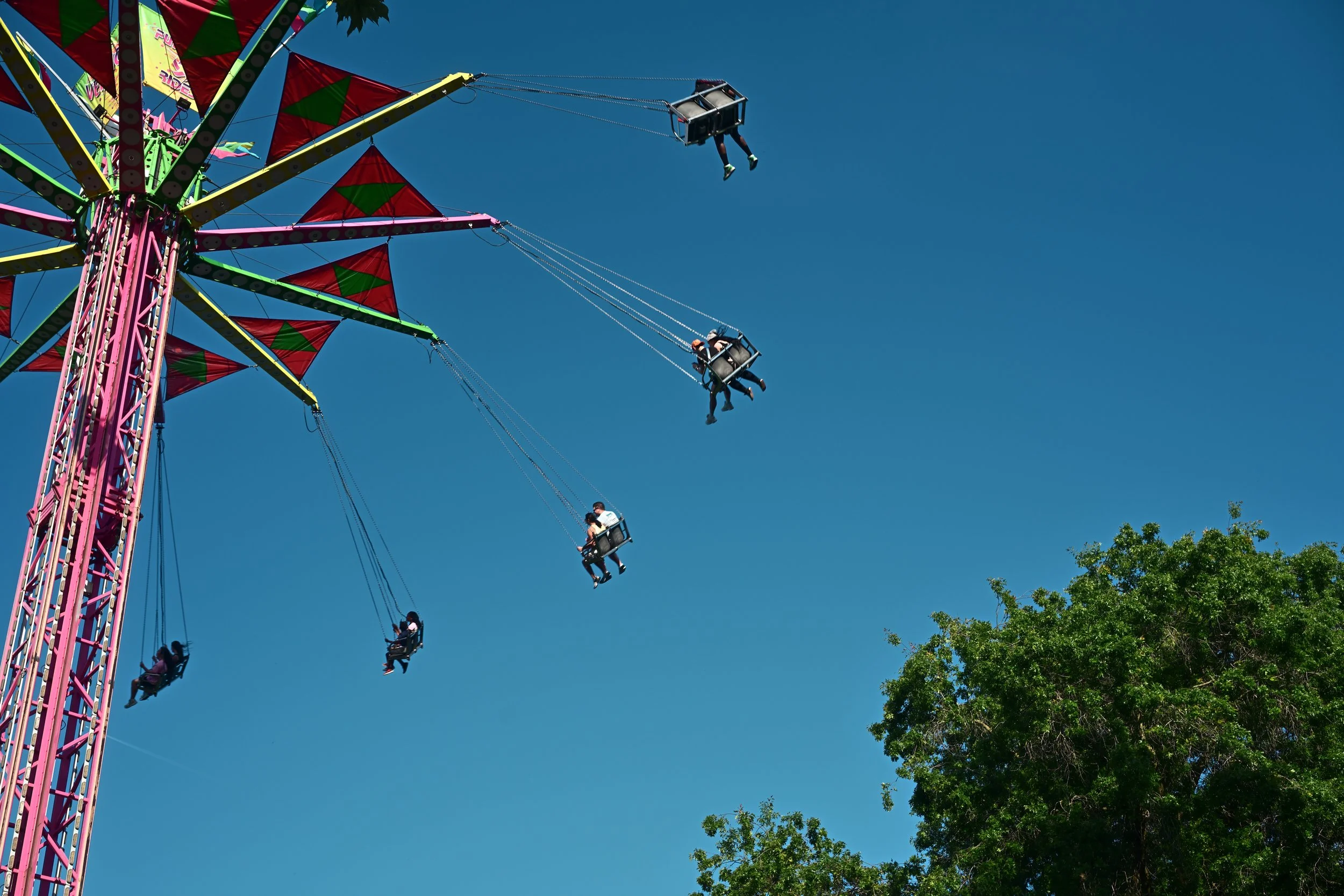 A colorful amusement park swing ride with children sitting in swings, swinging high into the clear blue sky with some green trees visible at the bottom right corner.