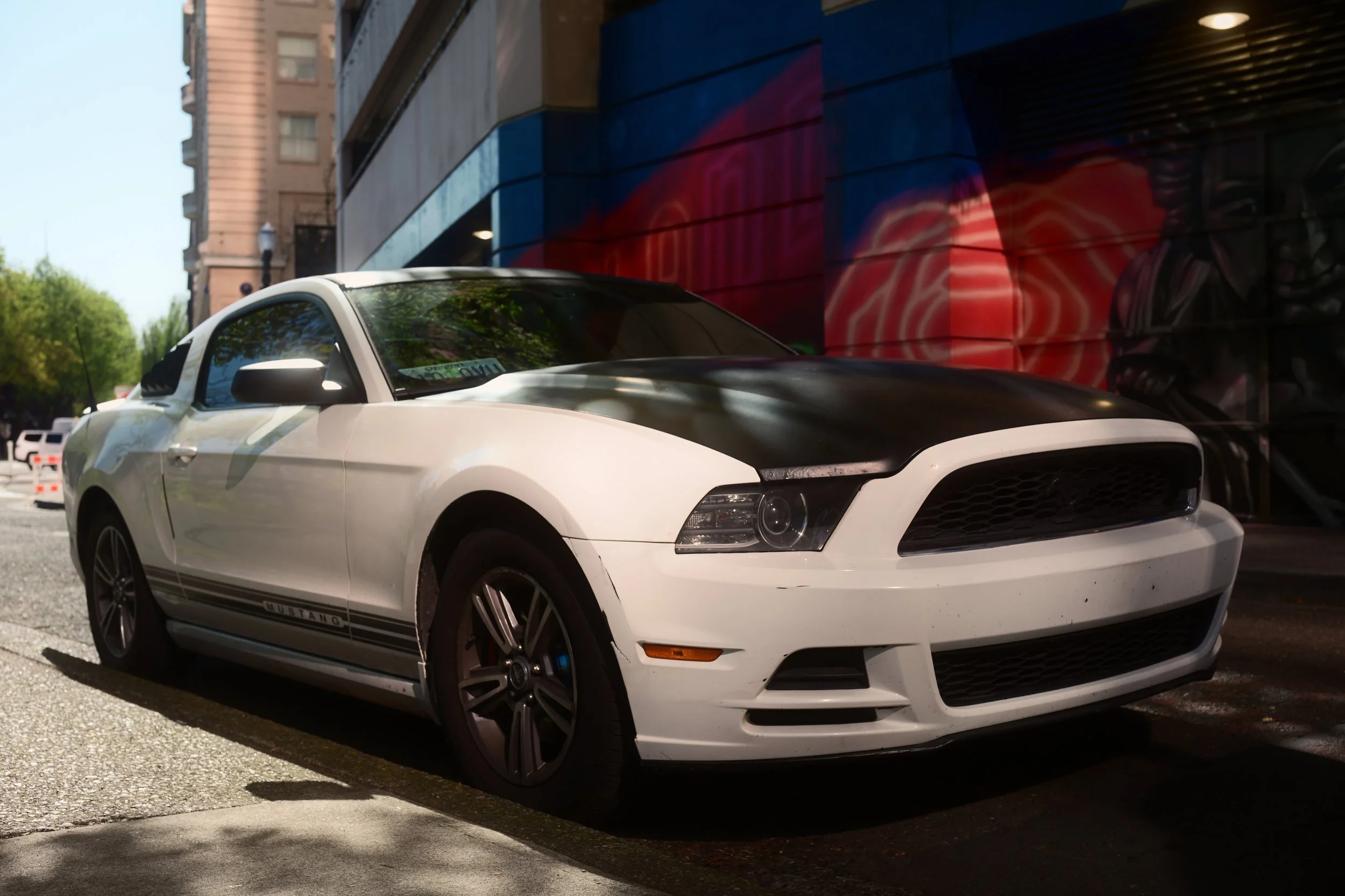 A white Ford Mustang car parked on the street next to a building with colorful mural art on its wall.