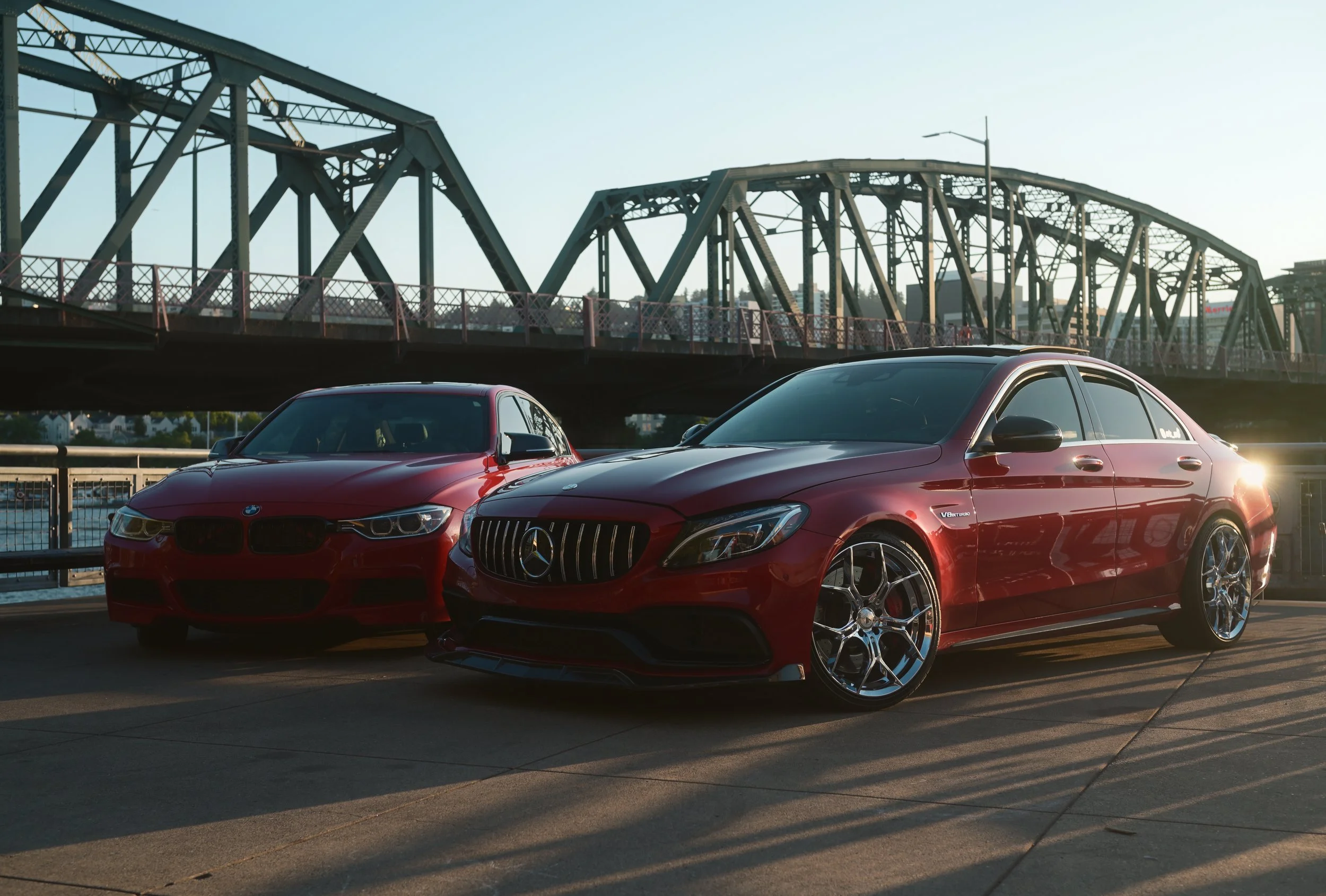 Two red luxury cars parked on a rooftop parking lot with a metal bridge structure in the background during sunset