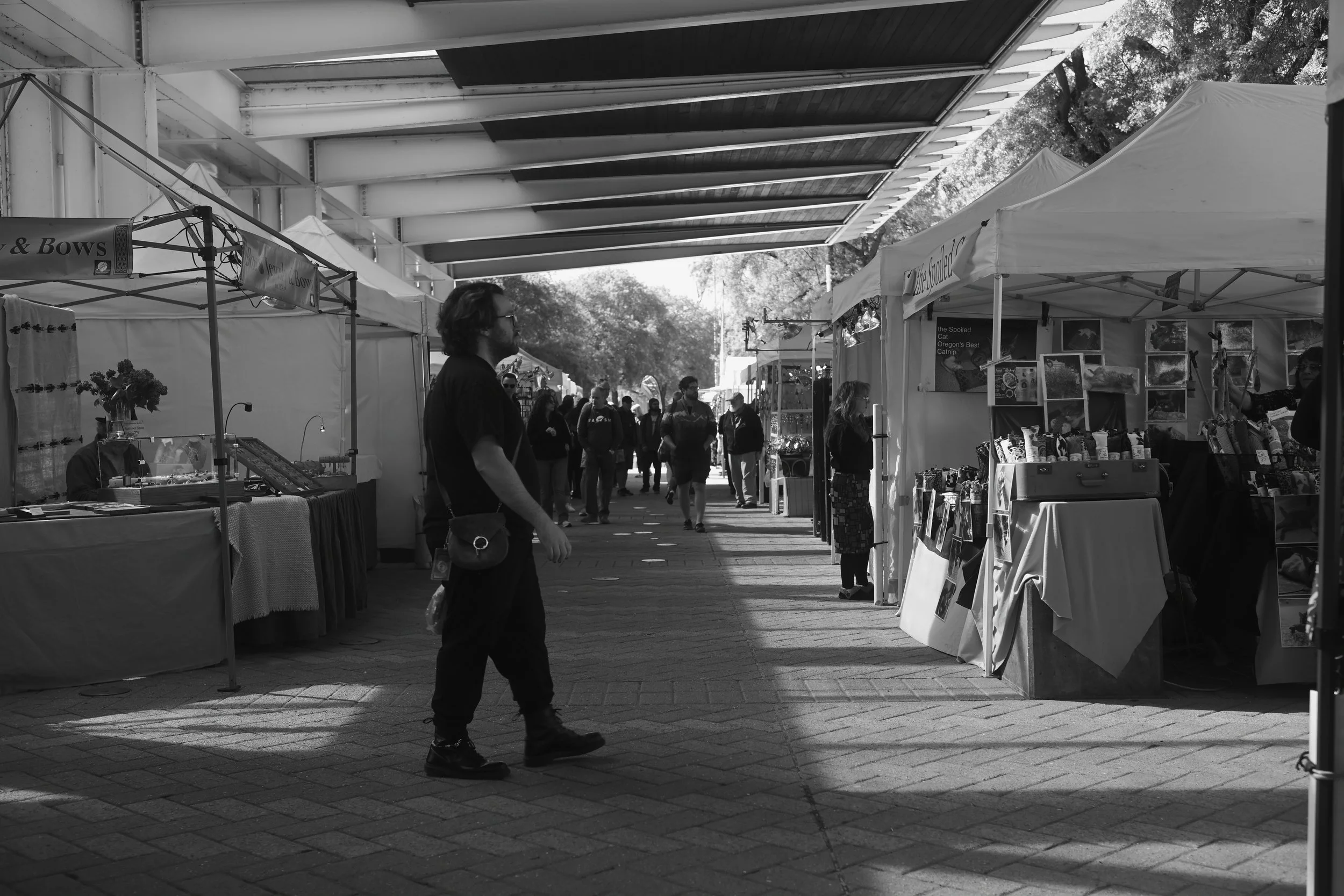 Black and white photo of an outdoor market with tents on either side, people walking and shopping, with trees and a sky overhead.