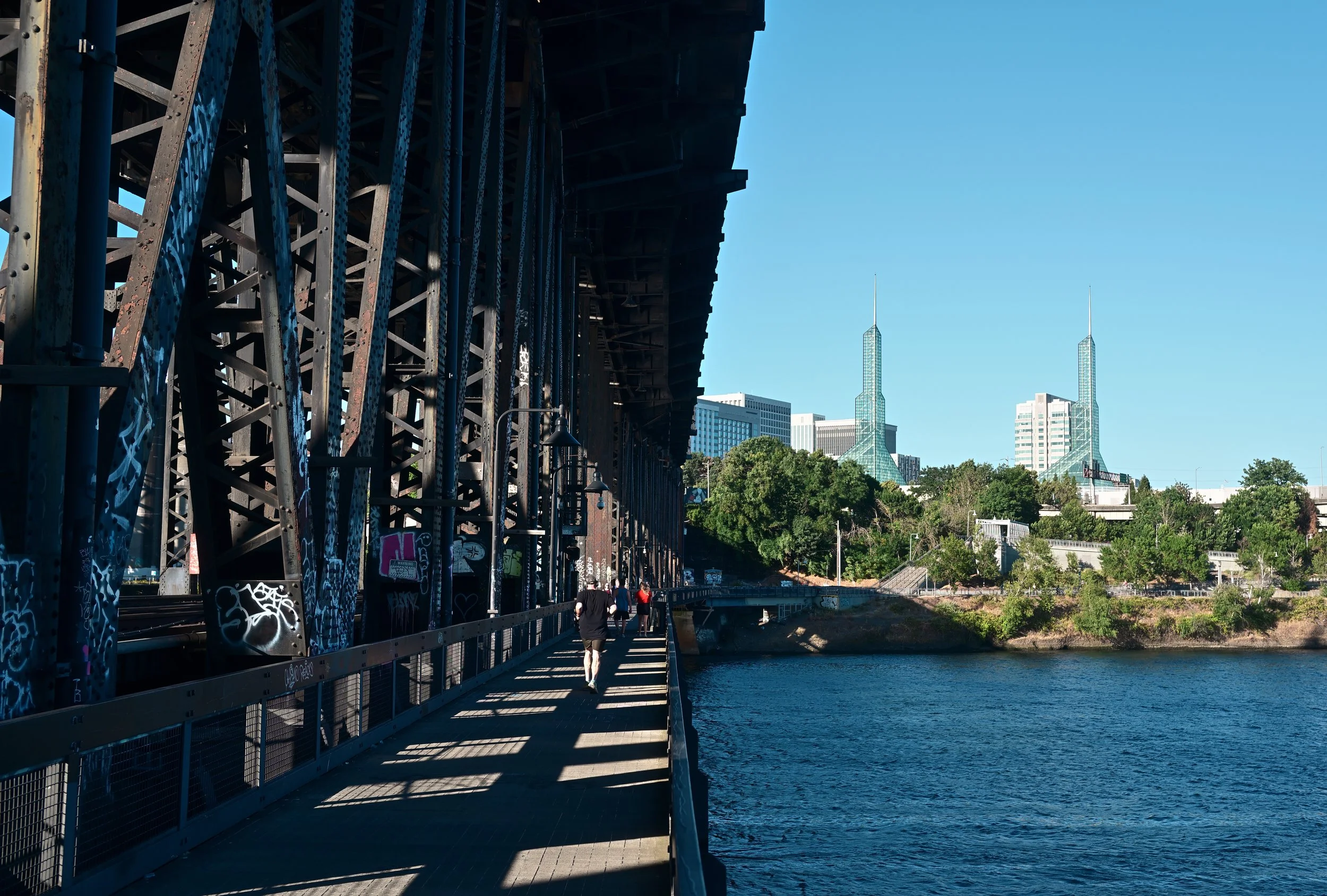 A footbridge over a river with a city skyline in the background, featuring two tall glass skyscrapers against a clear blue sky.