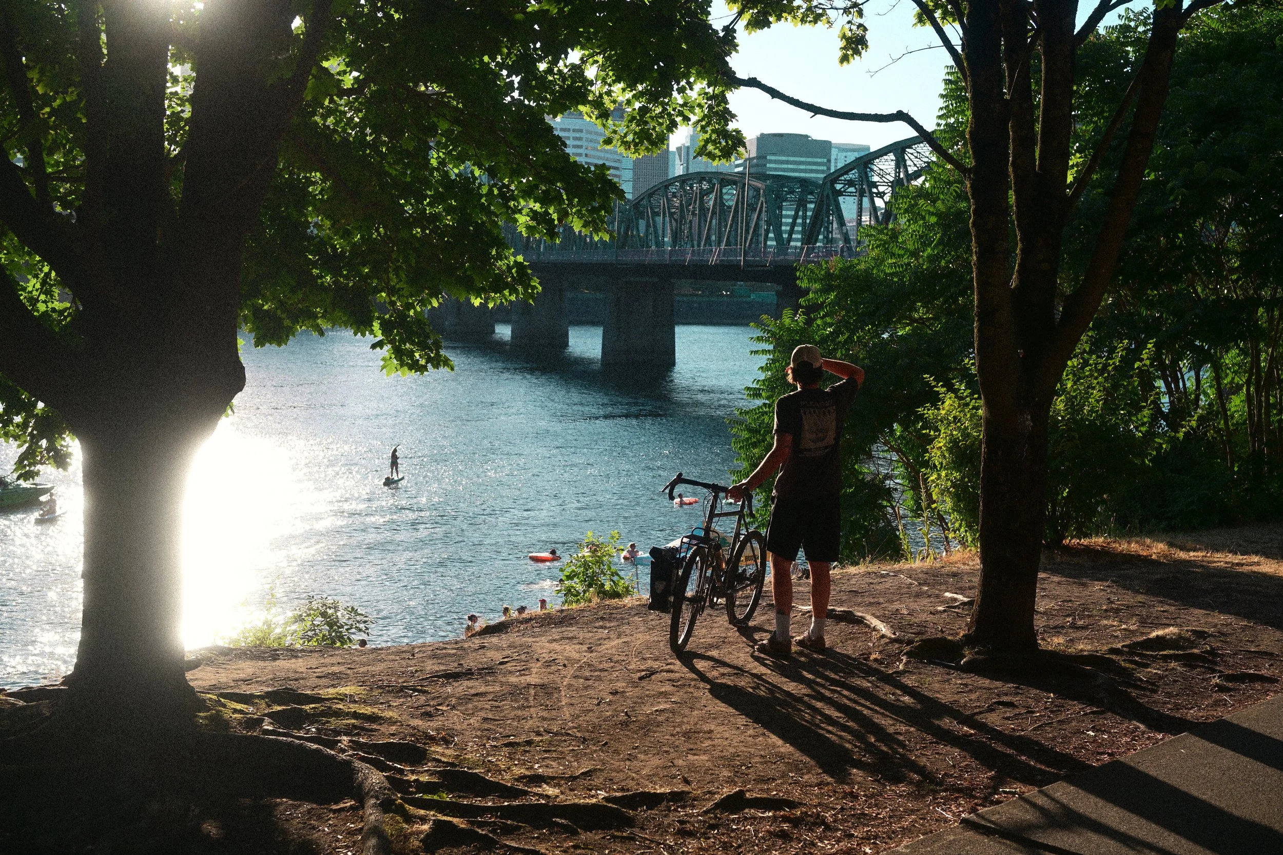 Cyclist with a bicycle overlooks swimmers on a river beneath a steel truss bridge, framed by trees and city buildings.