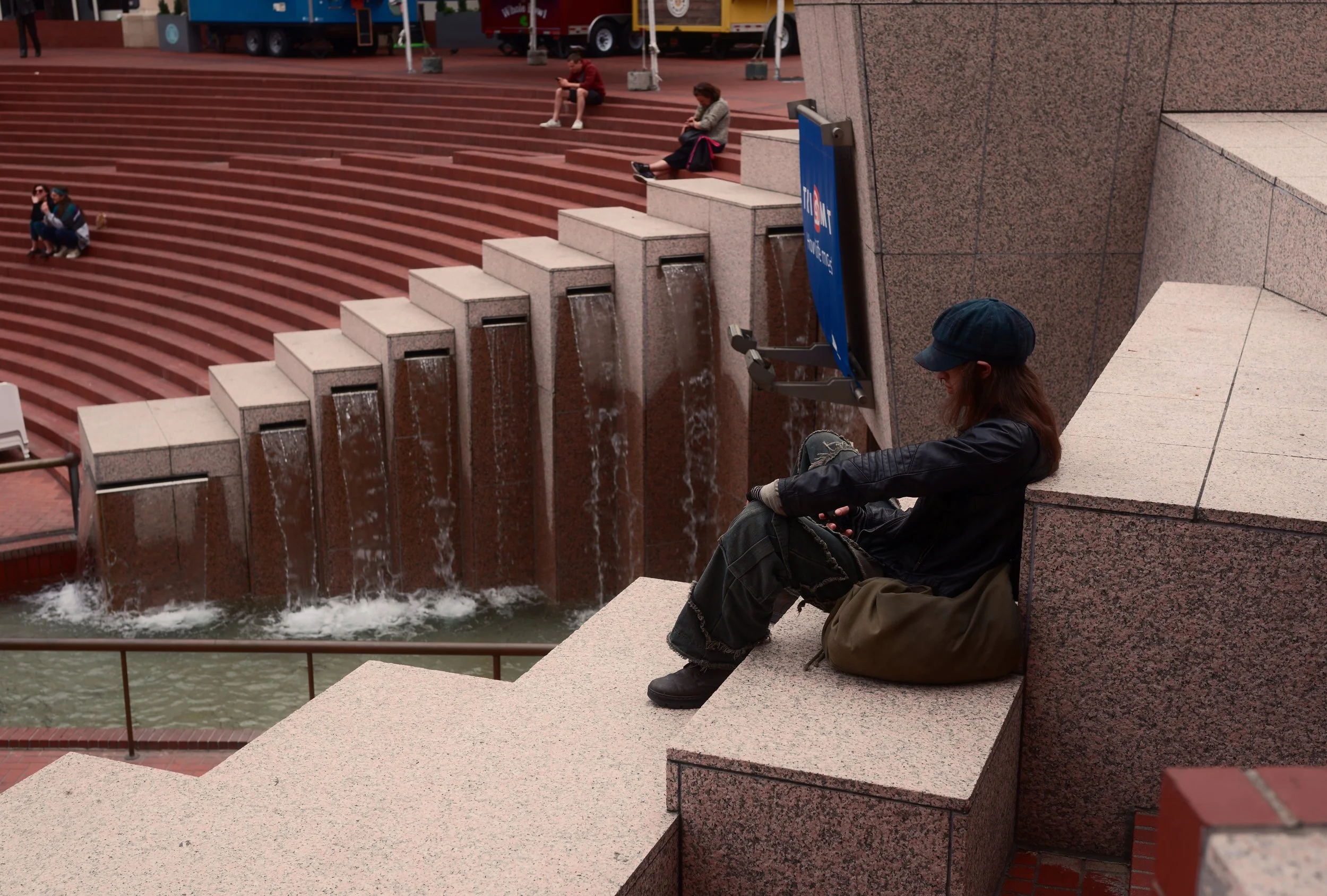 A person sitting on pink granite steps near a water fountain, wearing a dark jacket, jeans, and a cap, with a brown bag next to them. In the background, several people are sitting on the steps, some using their phones.