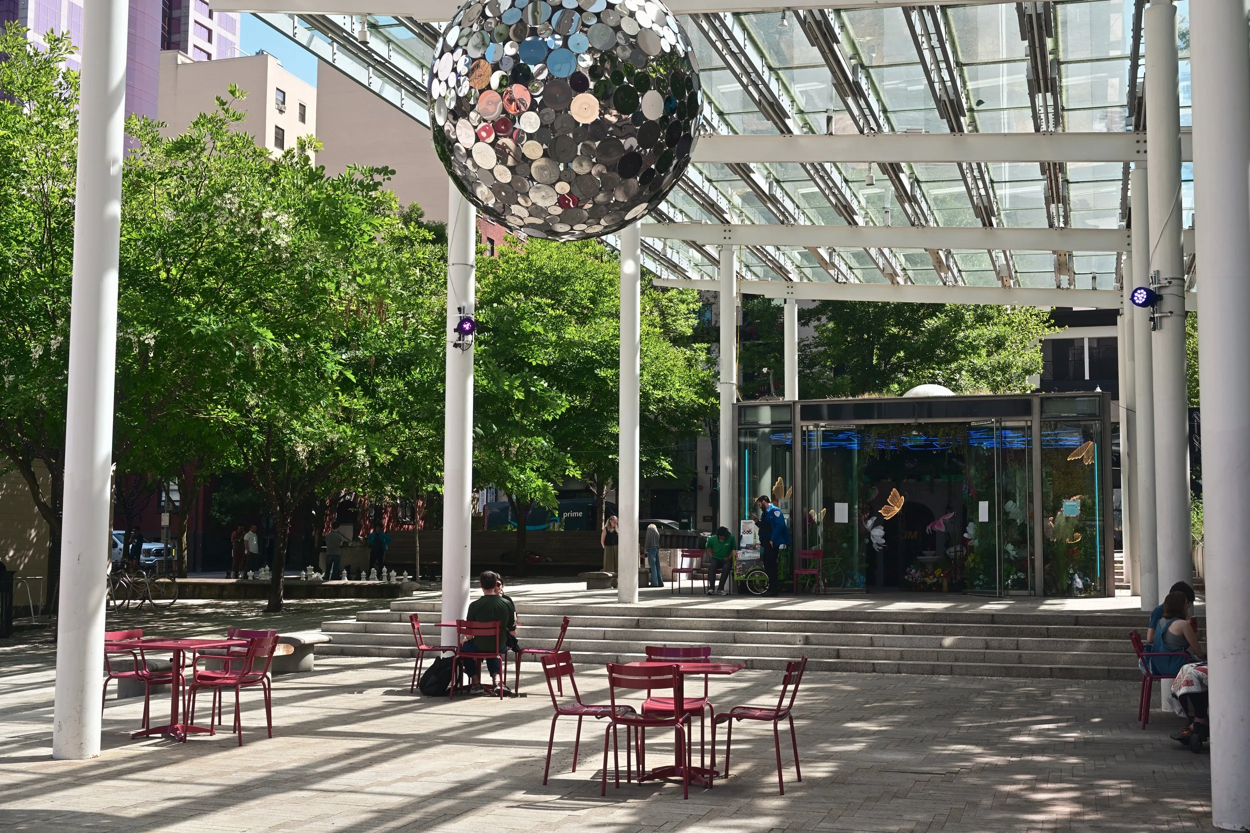 Outdoor urban plaza with trees, red tables and chairs, and people sitting and walking. A large spherical art installation made of mirrored discs hangs above. Modern glass and steel building structures are in the background.