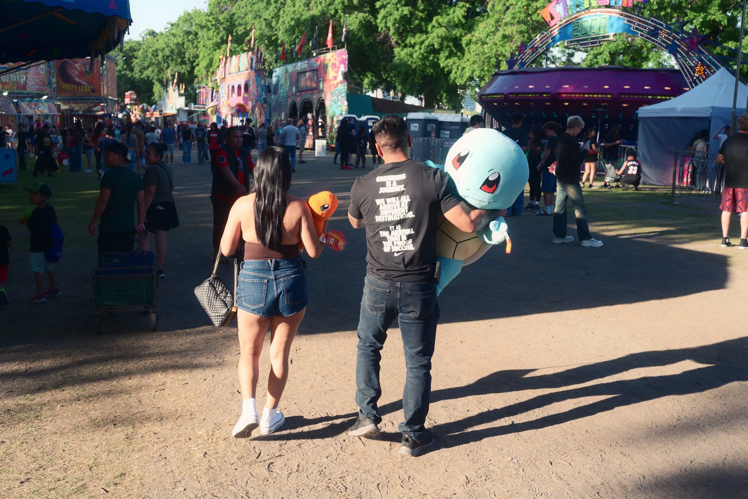 A young woman and a man are walking at a carnival, carrying plush Pokémon toys, including Squirtle. The carnival has rides and colorful booths, with many people enjoying the event on a sunny day.