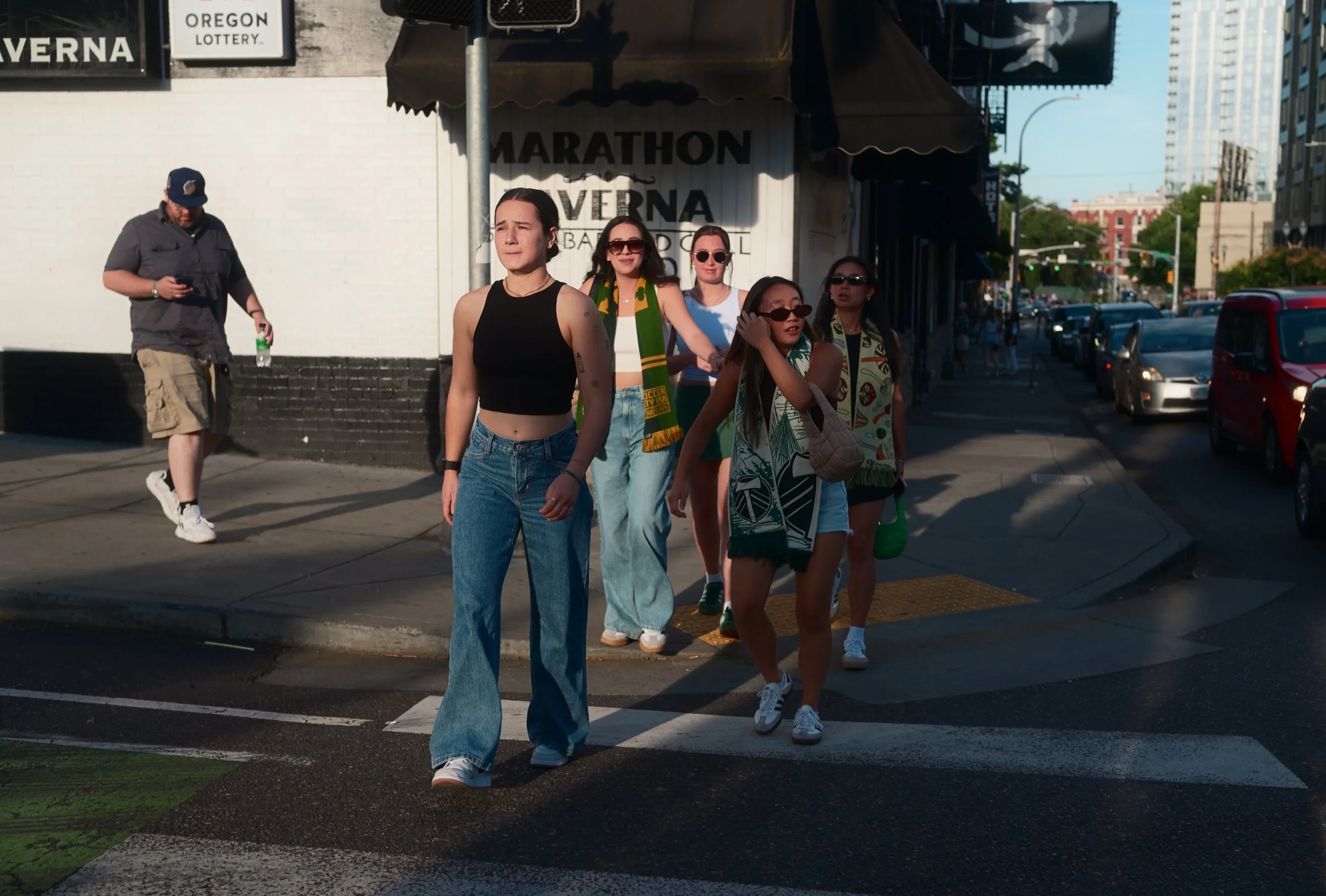 A group of five young women walking across a city street curb, one wearing a black crop top and blue jeans, others wearing sunglasses and casual summer clothing, with a man in the background looking at his phone, city buildings, parked cars, and stre