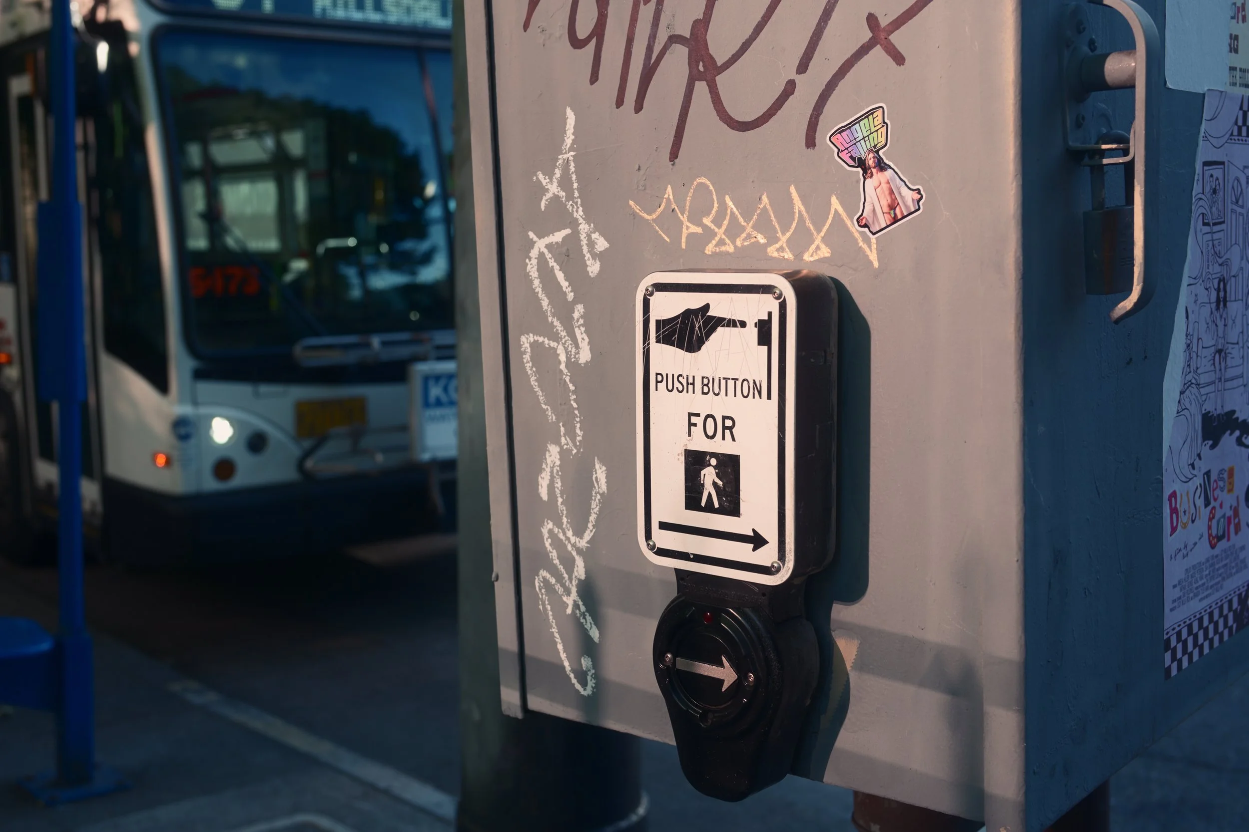Bus stop button with an arrow pointing to the right, mounted on a gray box covered with graffiti and stickers, with a bus in the background.
