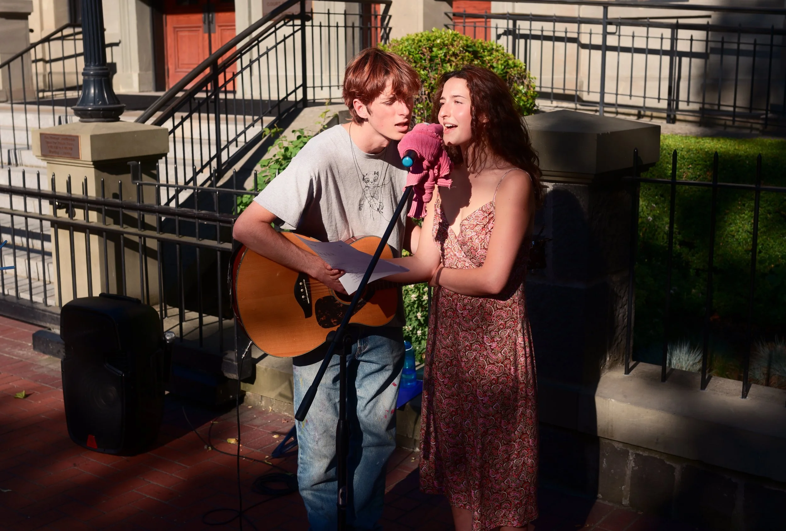 A young man with a guitar and a pink microphone cover singing into a microphone while a young woman in a patterned dress sings along outside.