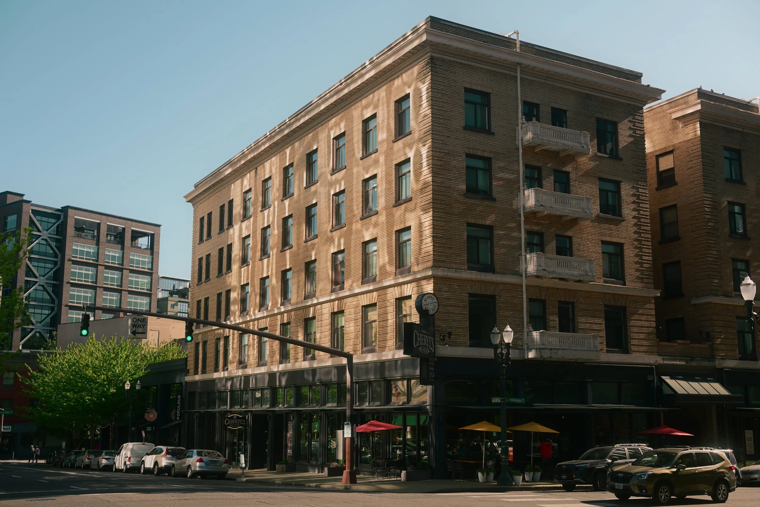 A city street scene featuring a multi-story brick building with balconies, large windows, and a ground-floor restaurant with outdoor seating. Cars are parked along the street, and a traffic light shows green.