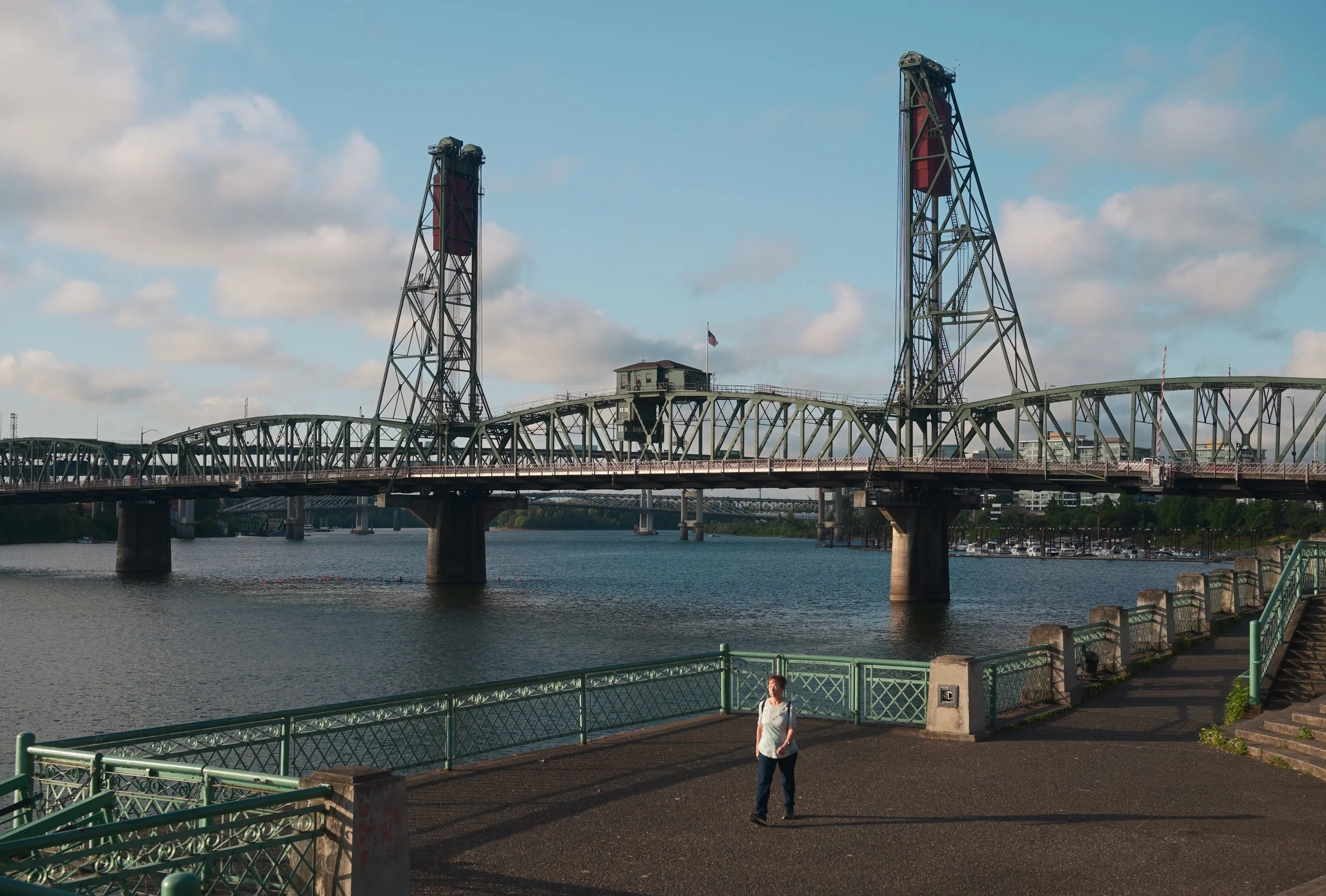 A scene of a bridge over a body of water with a walkway beside it, a woman walking and a person sitting nearby. In the background, there are two tall structures and a building with an American flag, under a partly cloudy sky.