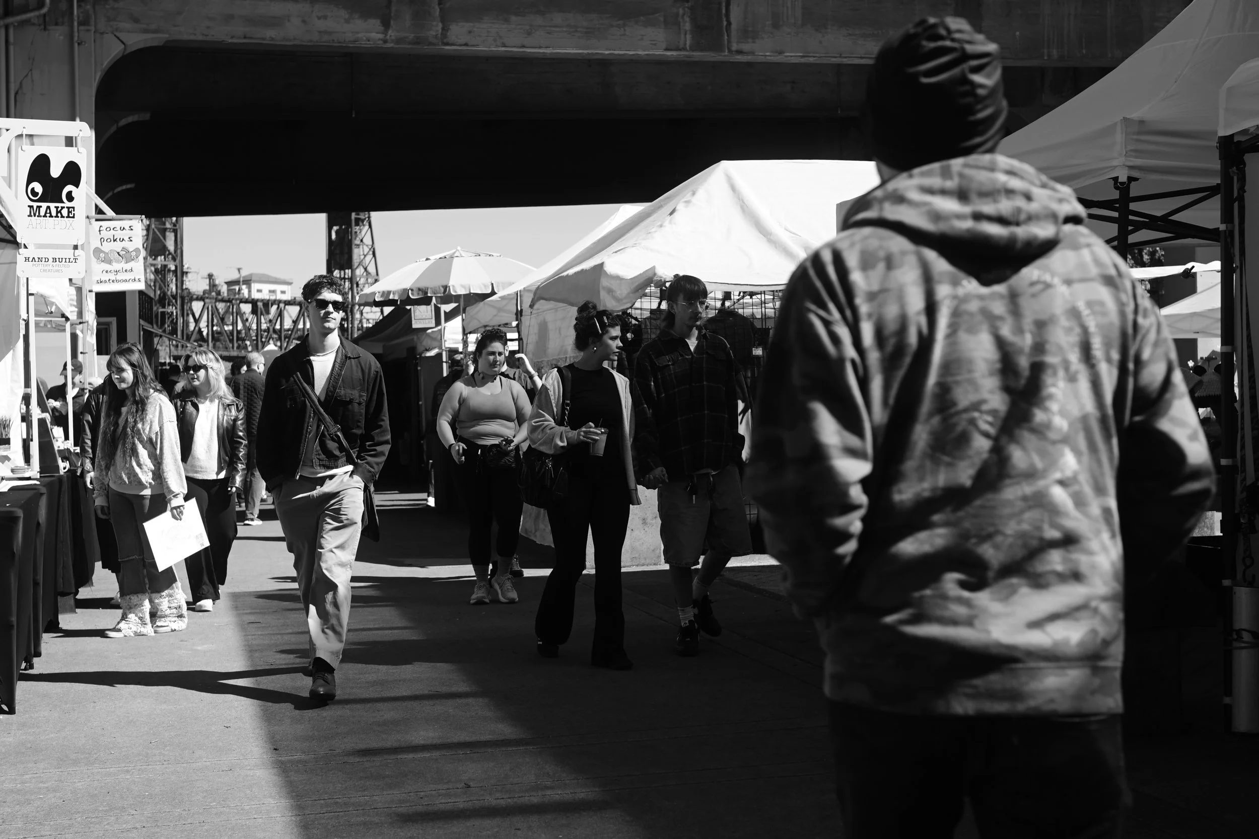 Black and white photo of people walking at an outdoor market or fair under a bridge, with tents and signs visible in the background.