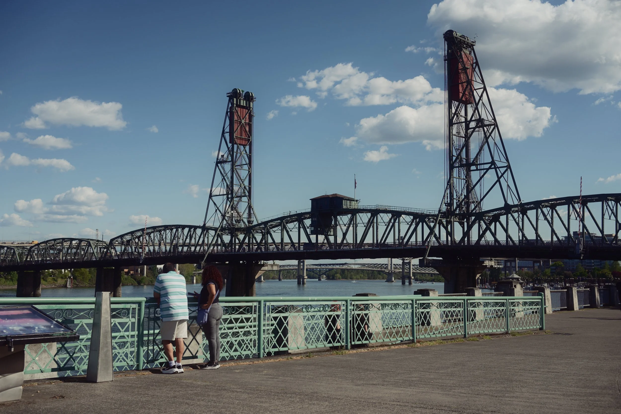 A bridge over a river with two tall oil derrick structures and a small building in the background under a partly cloudy sky, with two people standing by a green railing in the foreground.