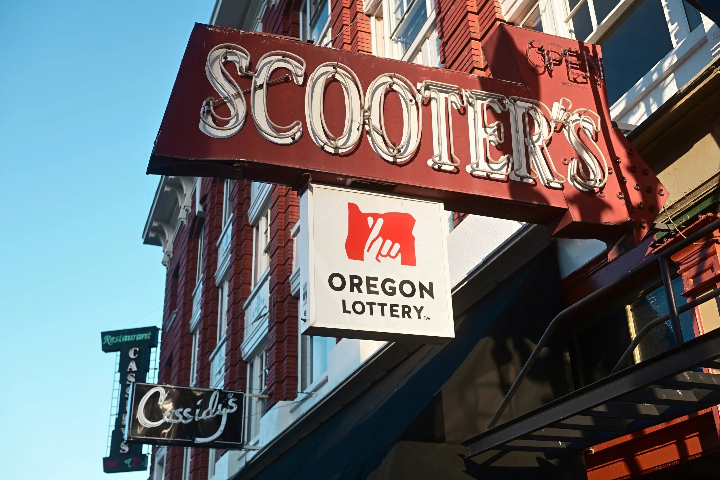 A red neon sign reading 'Open' and 'SCHOETERS' hanging above a storefront, with a smaller sign below indicating 'Oregon Lottery.' The building has a red brick facade with multiple windows, and nearby signage for a restaurant and casino can be seen al