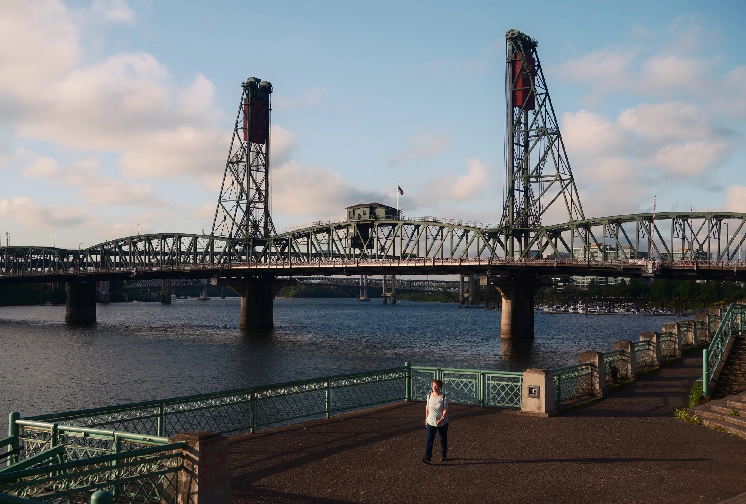A woman standing on a riverside promenade with green railing, overlooking a bridge crossing a body of water, with a blue sky and some clouds above.