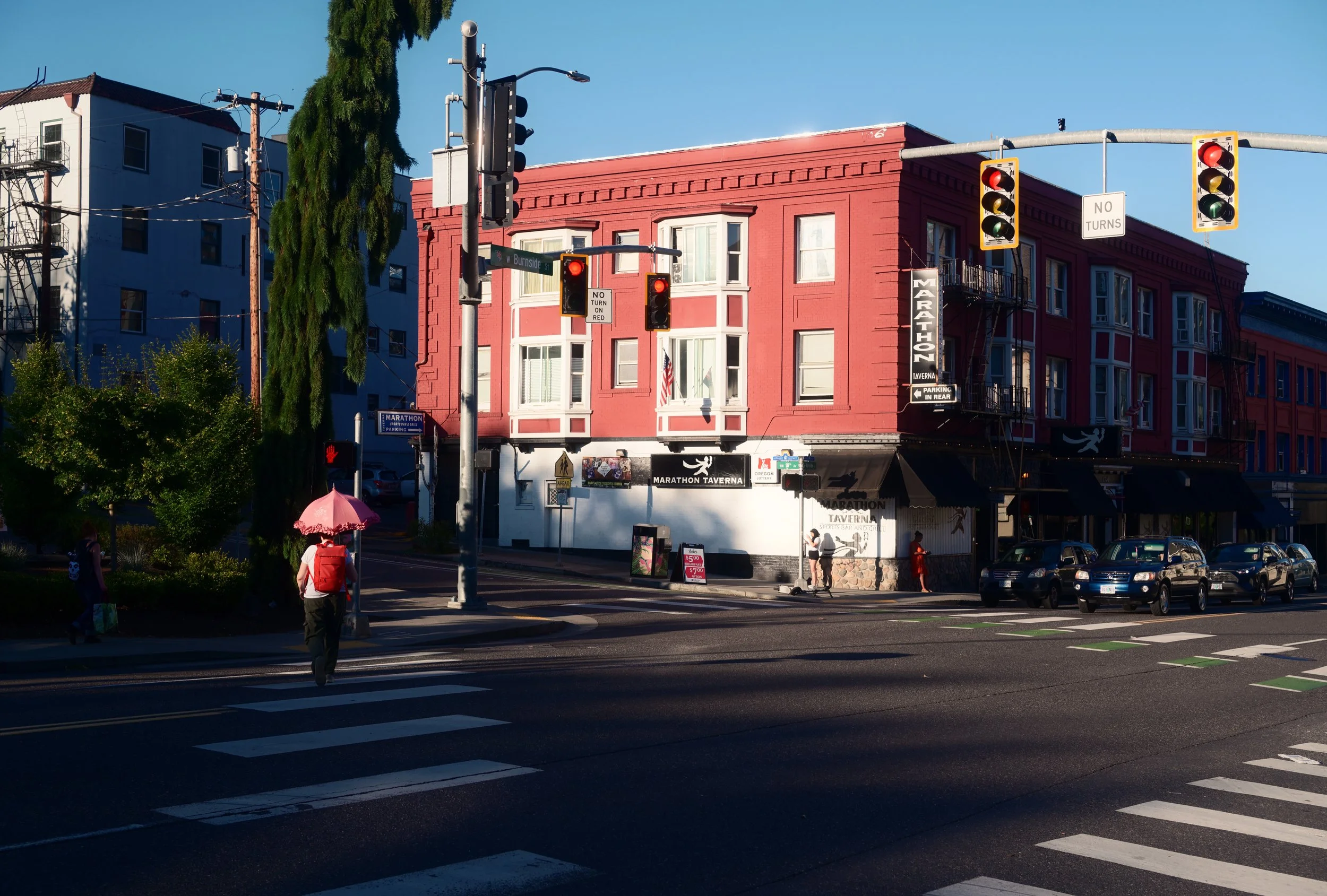 City street corner with red traffic lights, pedestrians crossing, and a pink building labeled 'Marathon Tavern' on a sunny day.