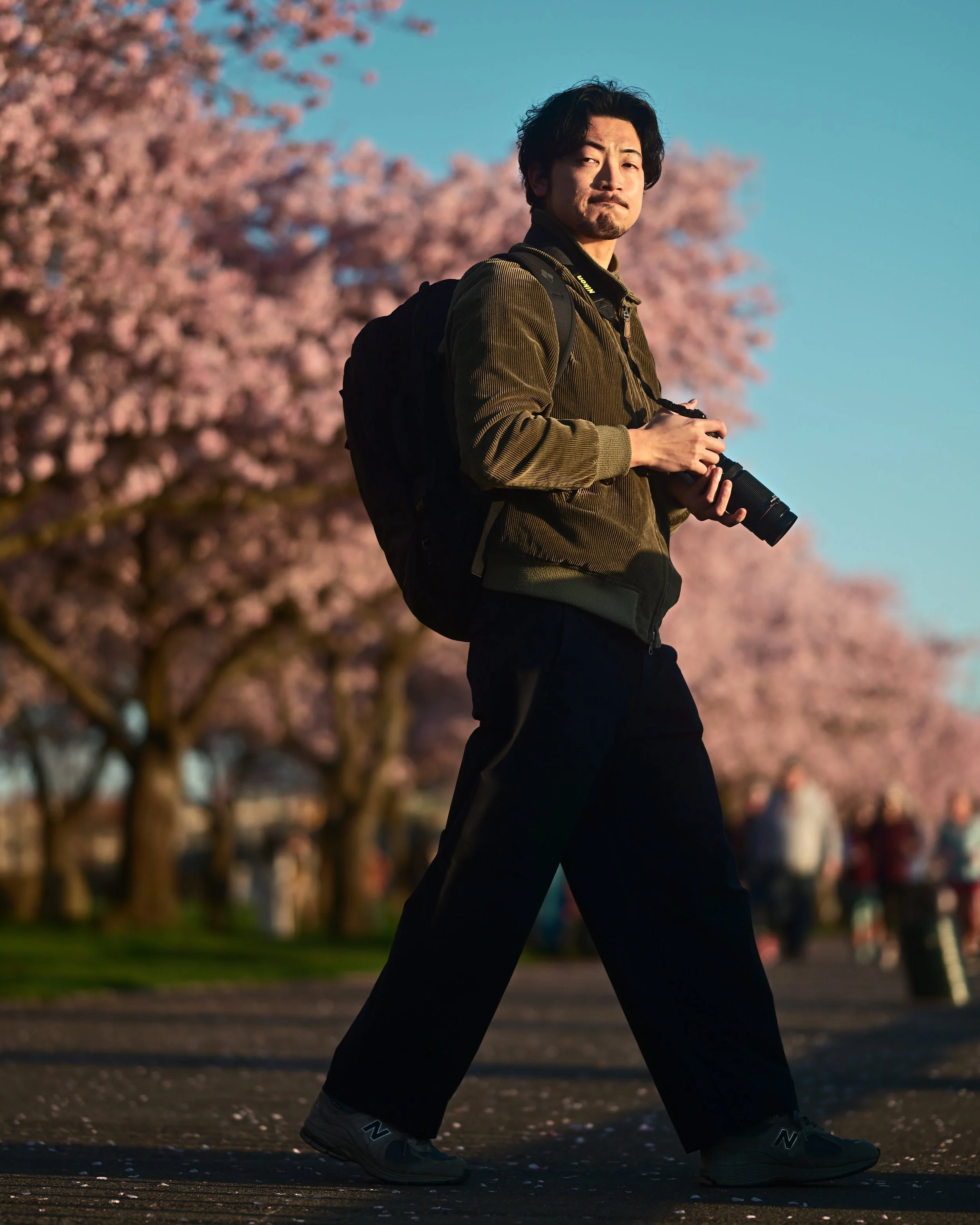 A man walking outdoors during cherry blossom season, holding a camera, wearing a backpack, dark pants, sneakers, and a brown jacket, with pink cherry blossoms and a clear blue sky in the background.