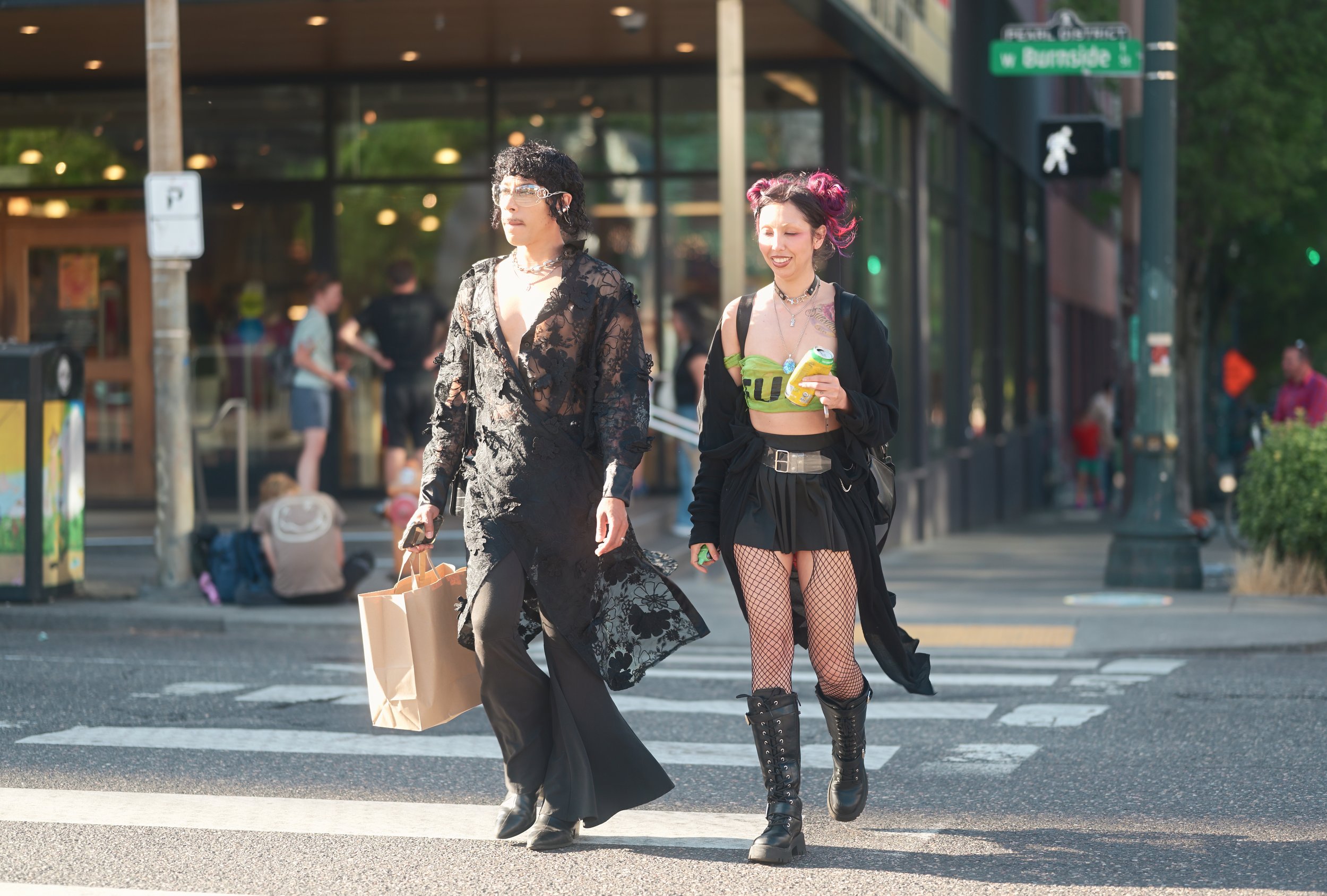 Two women crossing the street at a crosswalk in an urban area, one holding shopping bags and the other holding a drink, both dressed in alternative fashion styles.