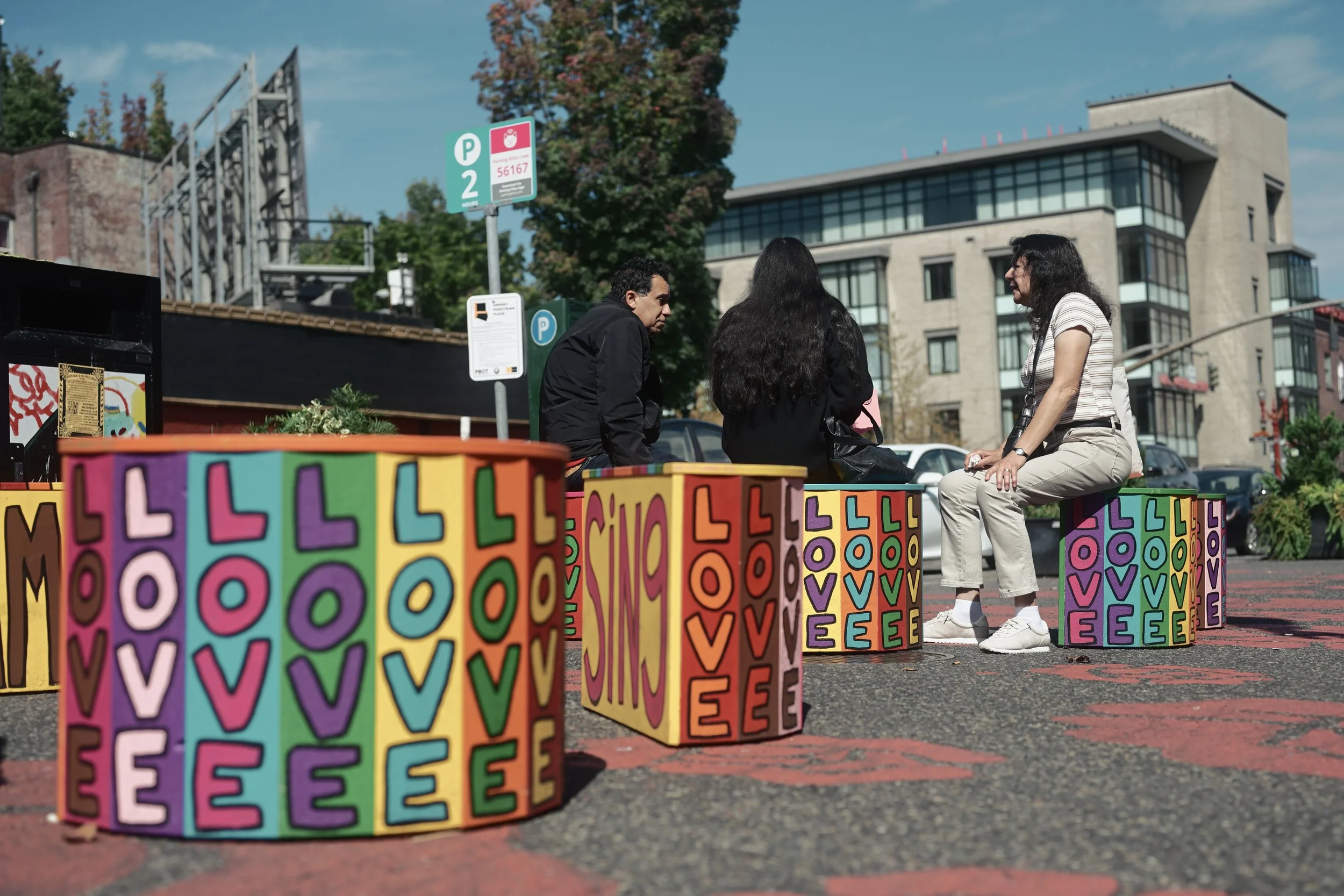 Three people sitting on a colorful ledge in an outdoor urban area with modern buildings and large trees in the background. The ledge is decorated with letters and colors.