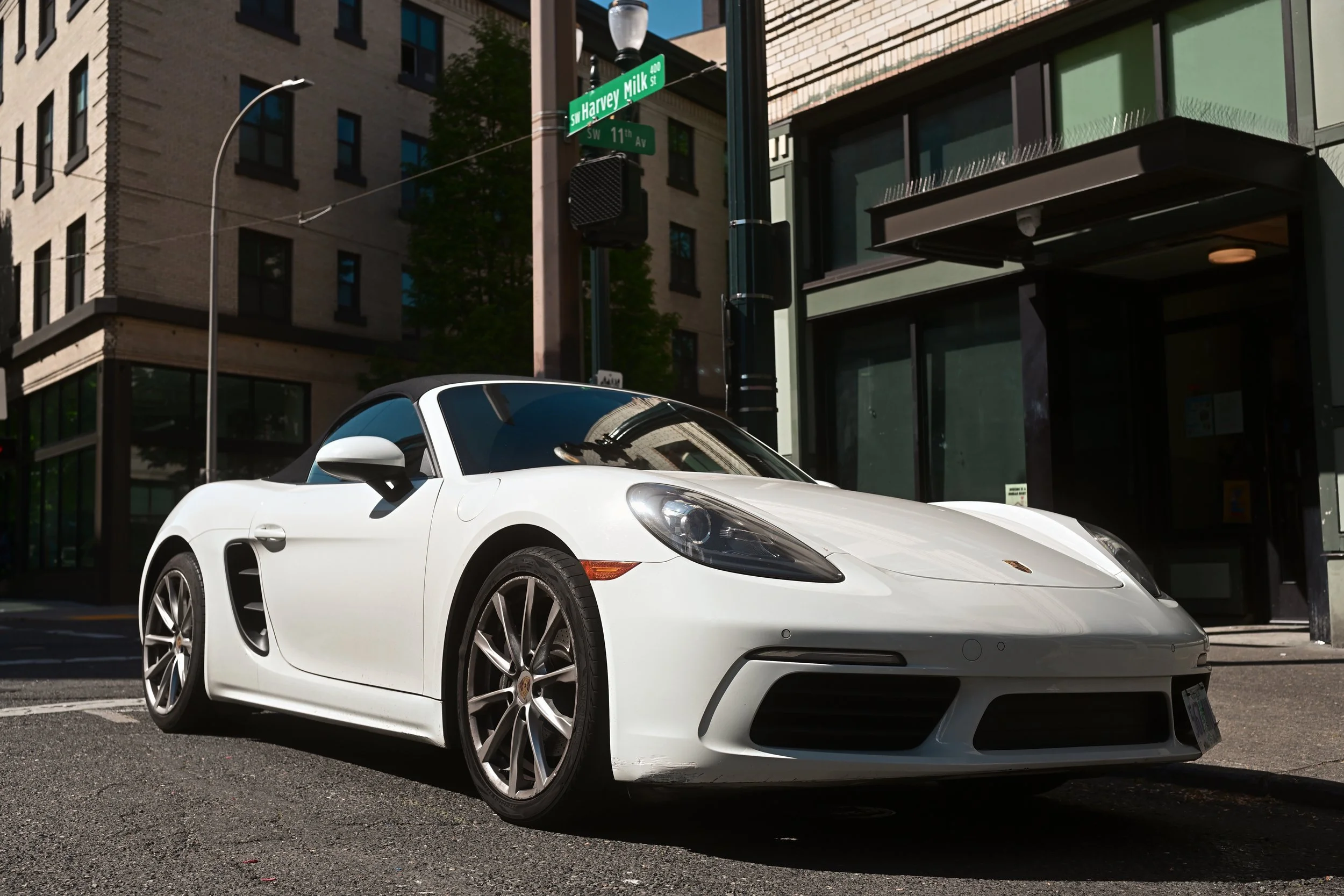 A white sports car parked on the street in front of a modern building with glass windows. Street signs at the corner indicate SW Harvey Milk St and SW 11th Ave. A street lamp and building facades are also visible.