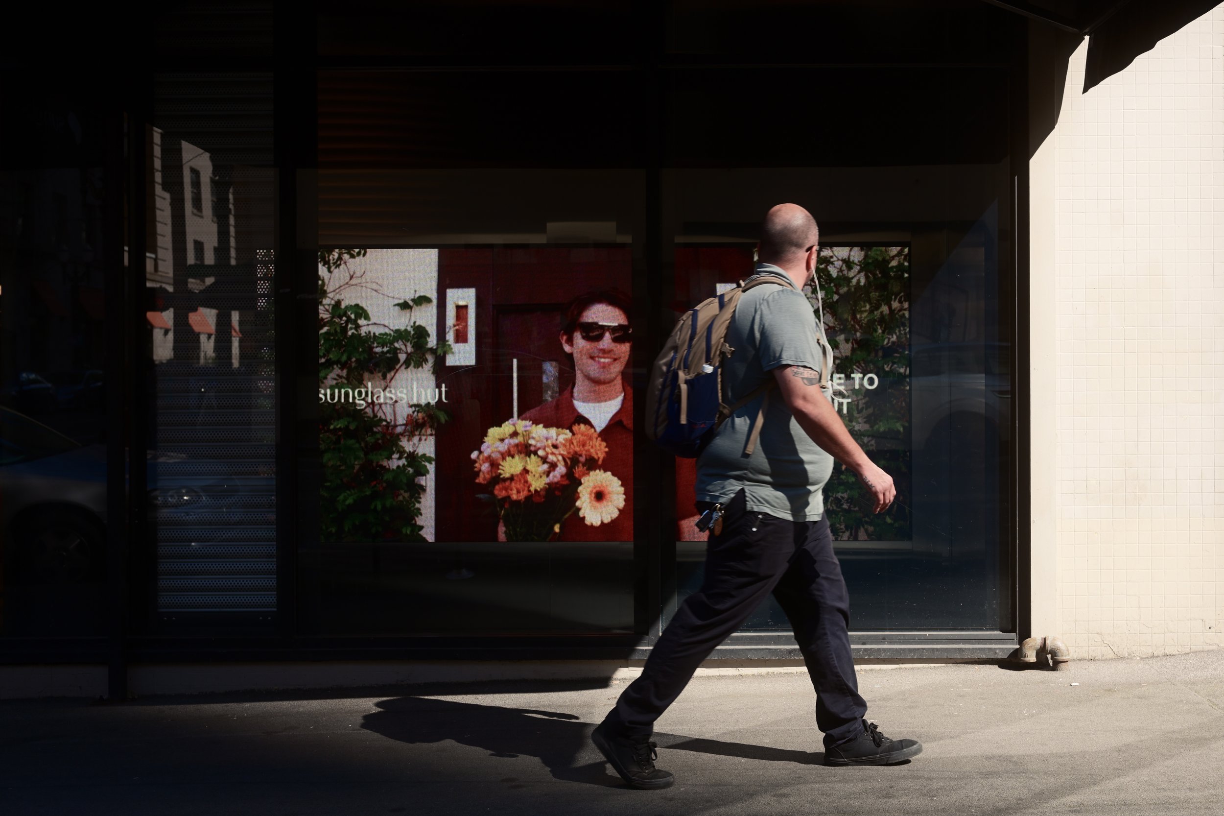 A man with a bald head and tattoos on his arm is walking past a store window that displays a portrait of a smiling man wearing sunglasses, holding a bouquet of flowers.