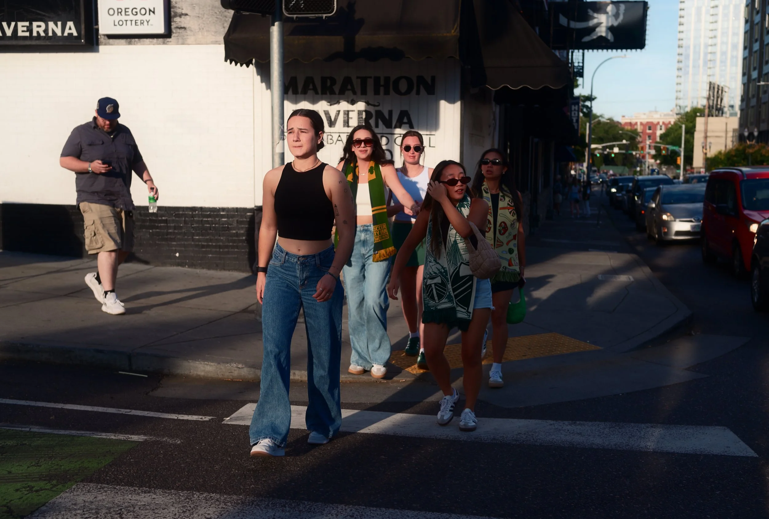 Group of six women crossing the street at a crosswalk in the sunlight, one woman in the front wearing a black sleeveless top and blue jeans, three women wearing sunglasses, and a man with a cap and cargo shorts in the background.