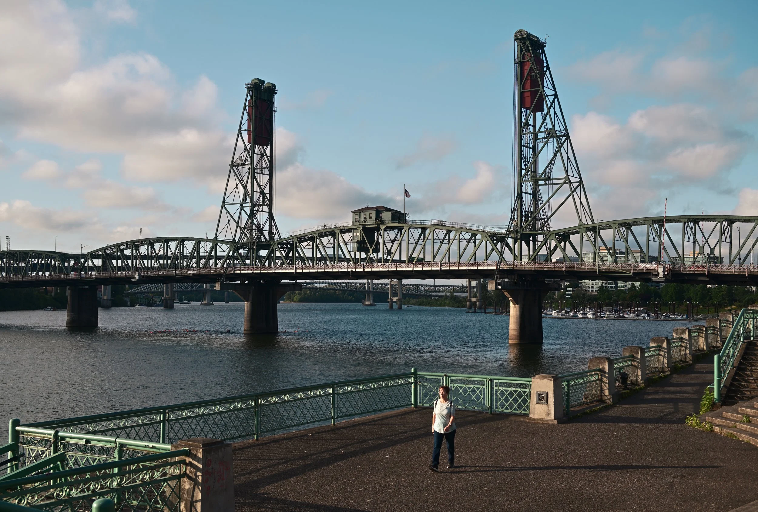 A river with a bridge overhead, two industrial-style towers, a building with an American flag, and a person walking along a waterfront walkway.