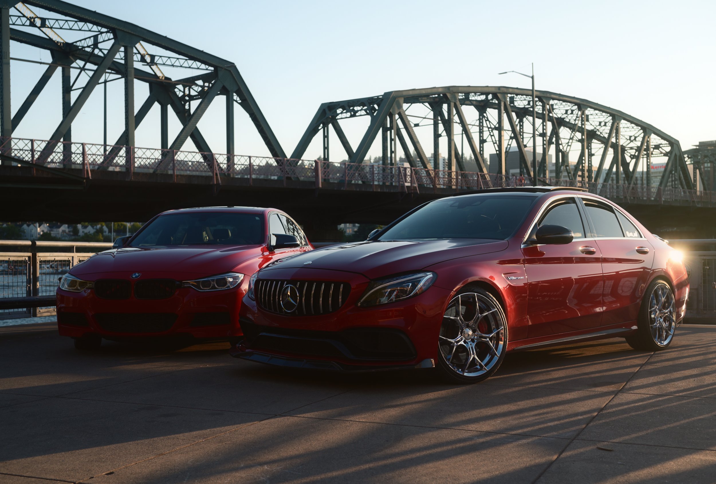 Red Mercedes-Benz and BMW cars parked on a city street with a bridge in the background during sunset.