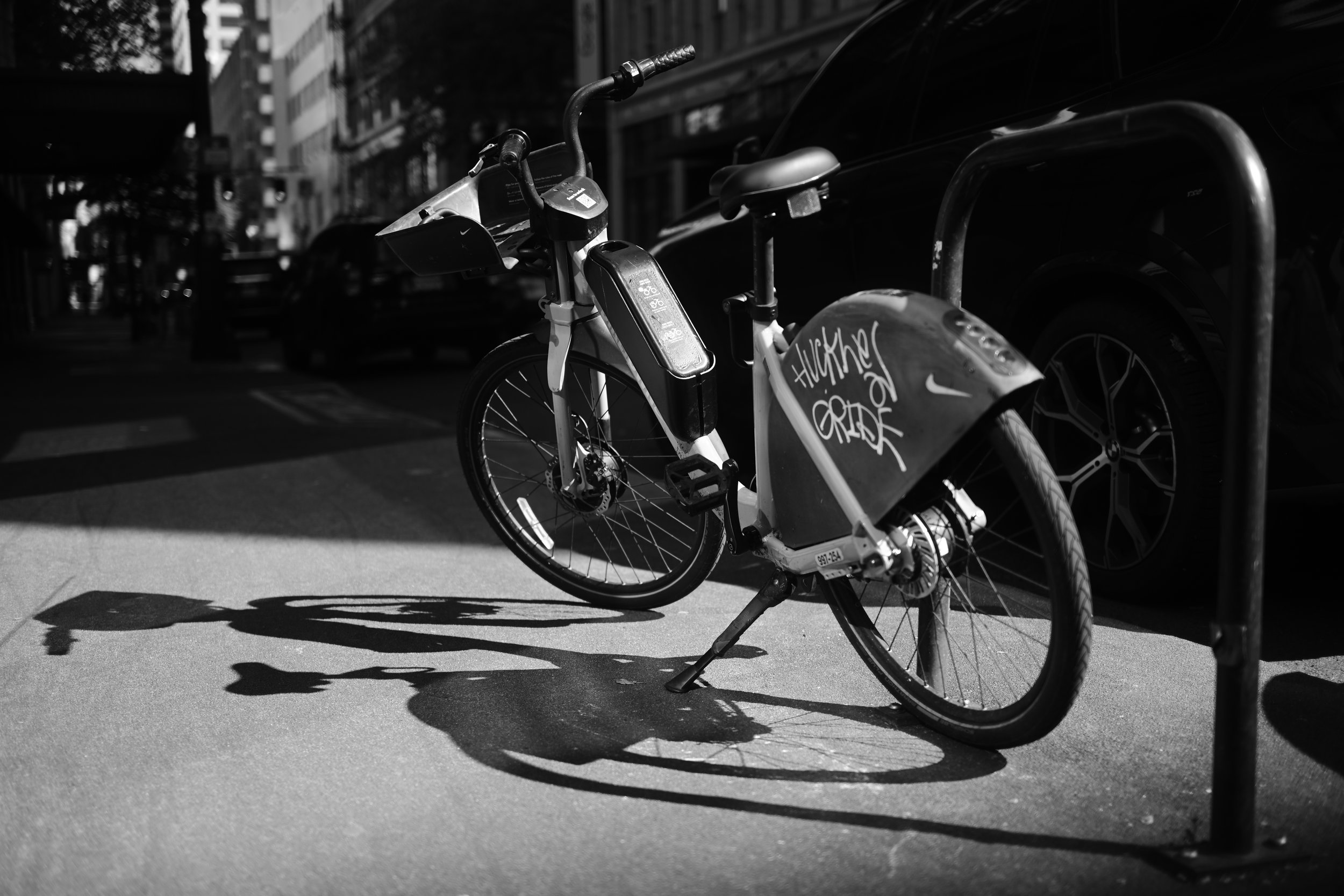 Black and white photo of a chained bicycle parked on a city sidewalk, casting a shadow on the ground.