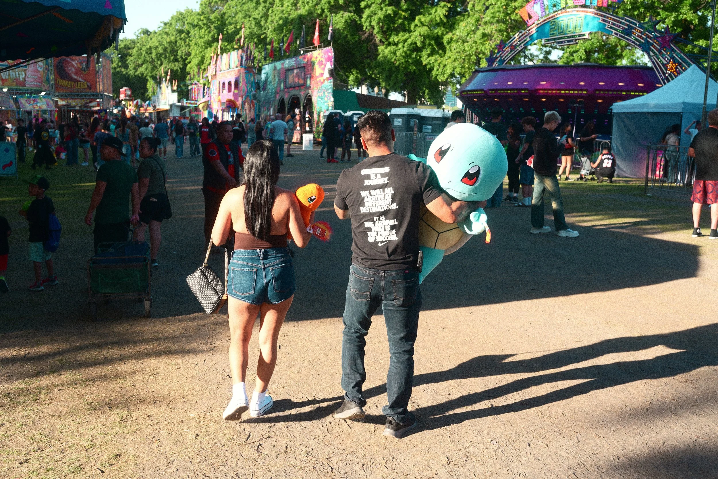Crowded outdoor festival with colorful carnival rides, decorated booths, and a person carrying a large blue alien mascot plush.