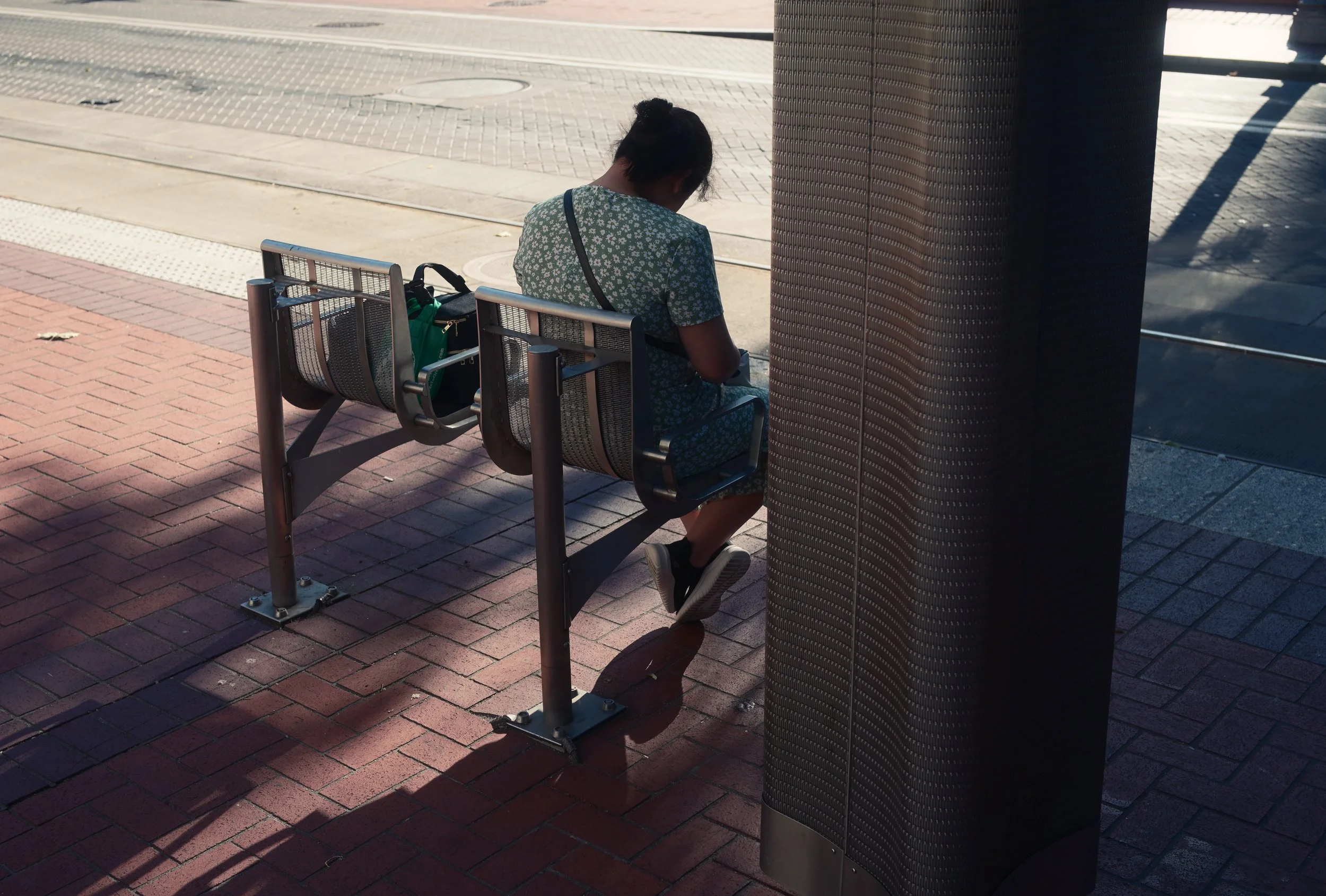 A woman with her hair tied back, sitting on a metal bench at a bus stop, looking at her phone. She has a green bag placed on the end of the bench next to her. The bus stop is on a sidewalk with a brick surface, and a textured dark column is nearby. T