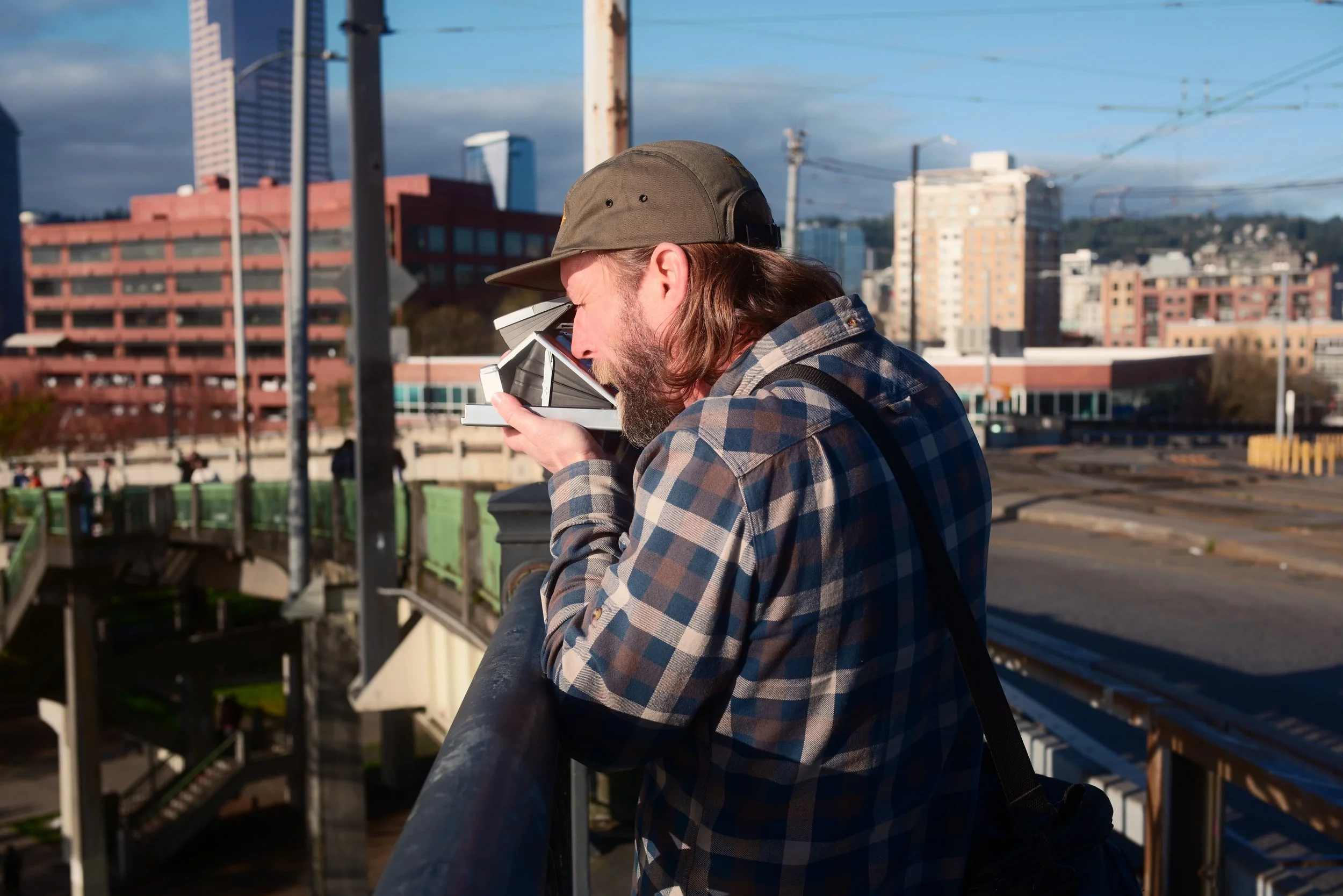 Man with long hair and beard wearing a baseball cap and plaid shirt looking through binoculars on a city bridge with buildings and power lines in the background.