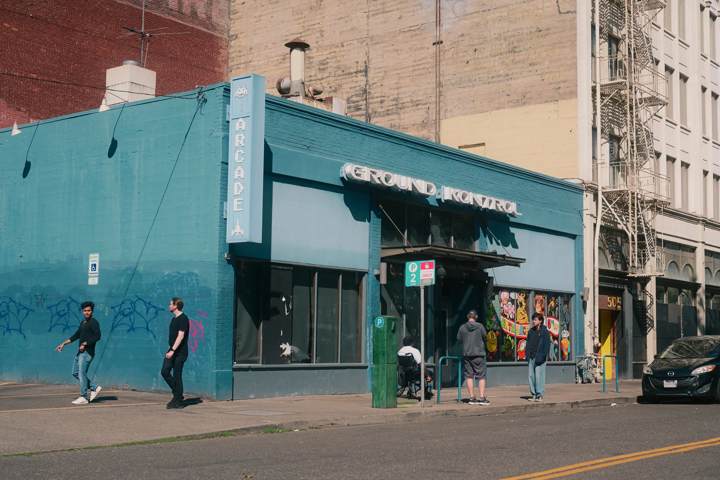 City street scene with people walking past a blue building with a pixel art arcade sign. The building has graffiti on the lower part and colorful street art on the side. There are parked cars, a parking meter, and a fire escape on the adjacent building.