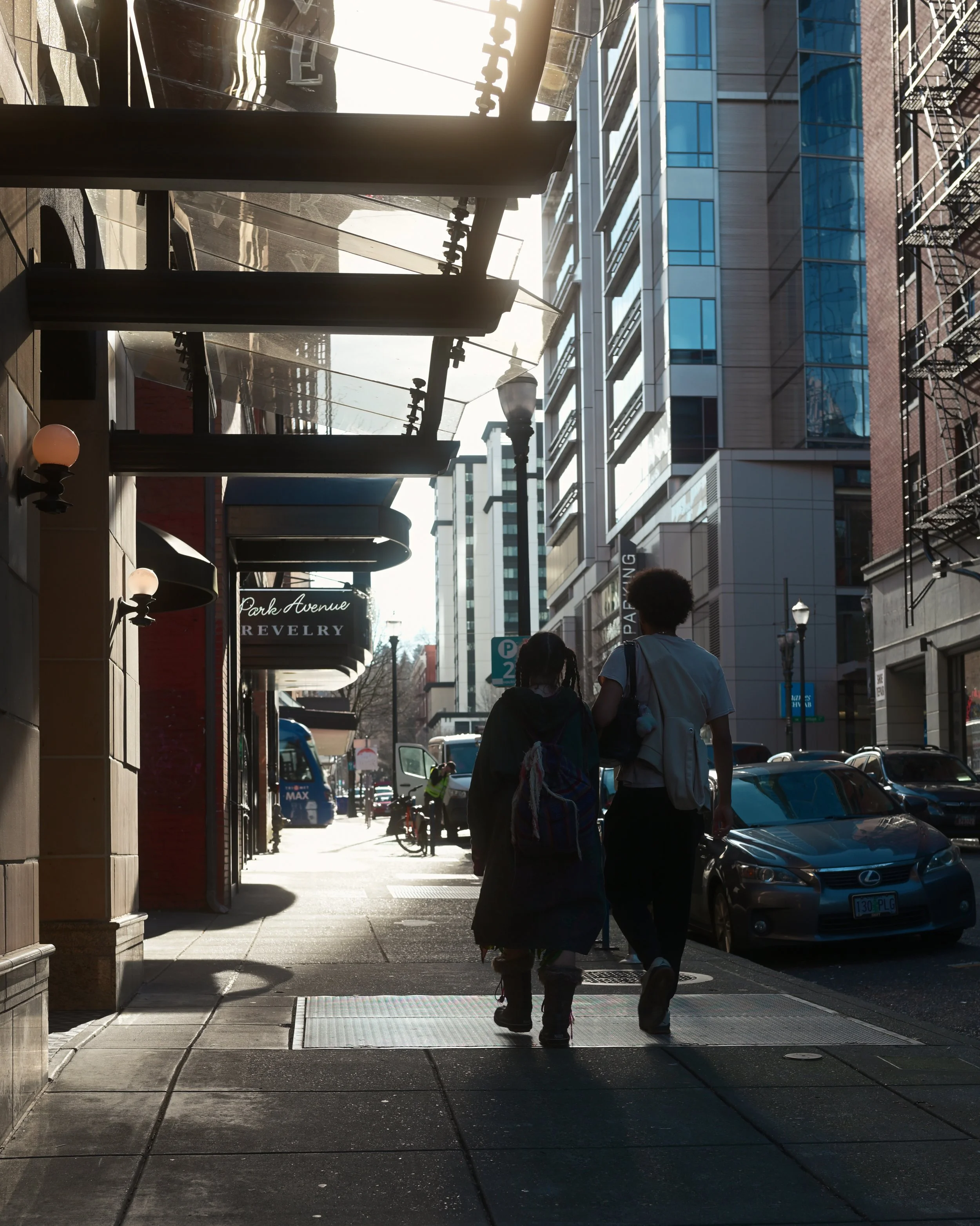People walking on a city sidewalk with modern buildings, cars parked along the street, and sunlight casting shadows. There are street lamps and signs including one that says 'Park Avenue'.
