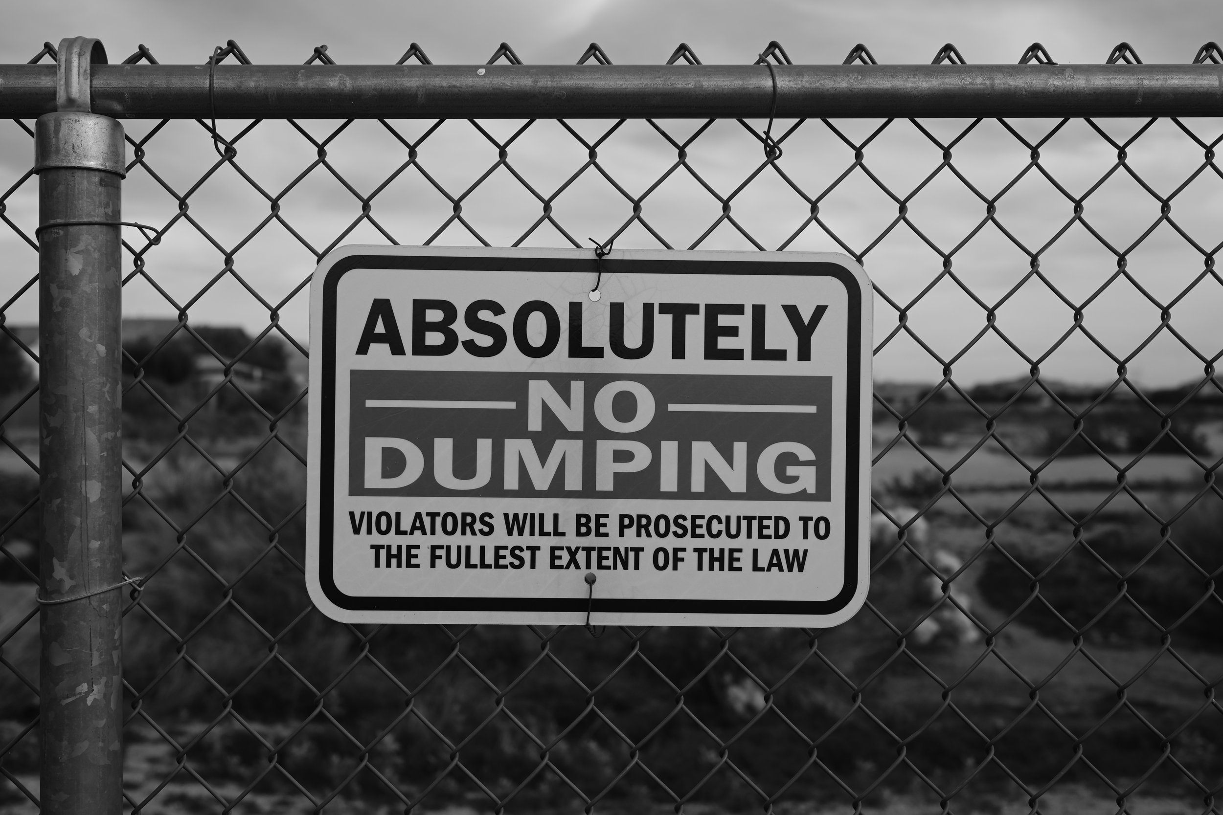 A chain-link fence with a sign that reads 'Absolutely No Dumping. Violators will be prosecuted to the fullest extent of the law.' in black and white.