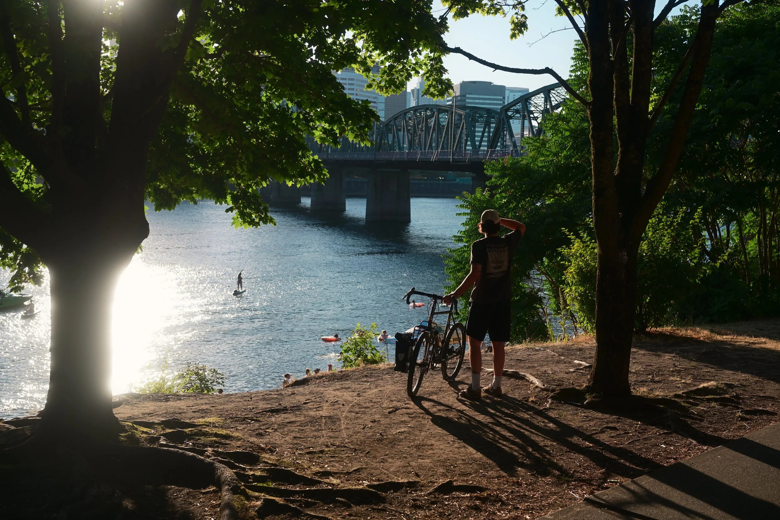 A person with a bicycle standing on a dirt path by a river, looking at people swimming and paddleboarding in the water, with a bridge and city buildings in the background, trees and sunlight creating a shaded foreground.