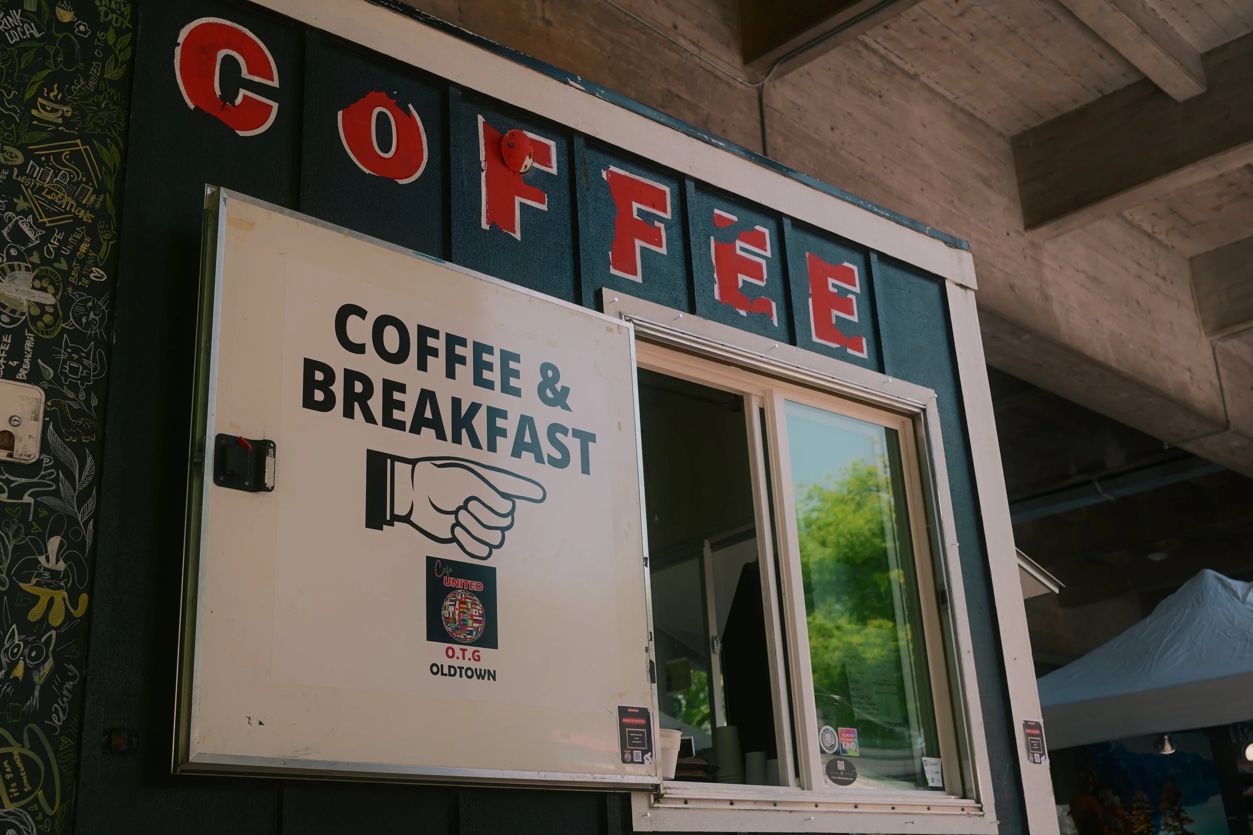View of a sign outside a coffee shop that reads 'Coffee' with red letters on a black background, and a smaller sign below that advertises coffee and breakfast, with a graphic of a hand pointing and the words 'Coffee & Breakfast'.